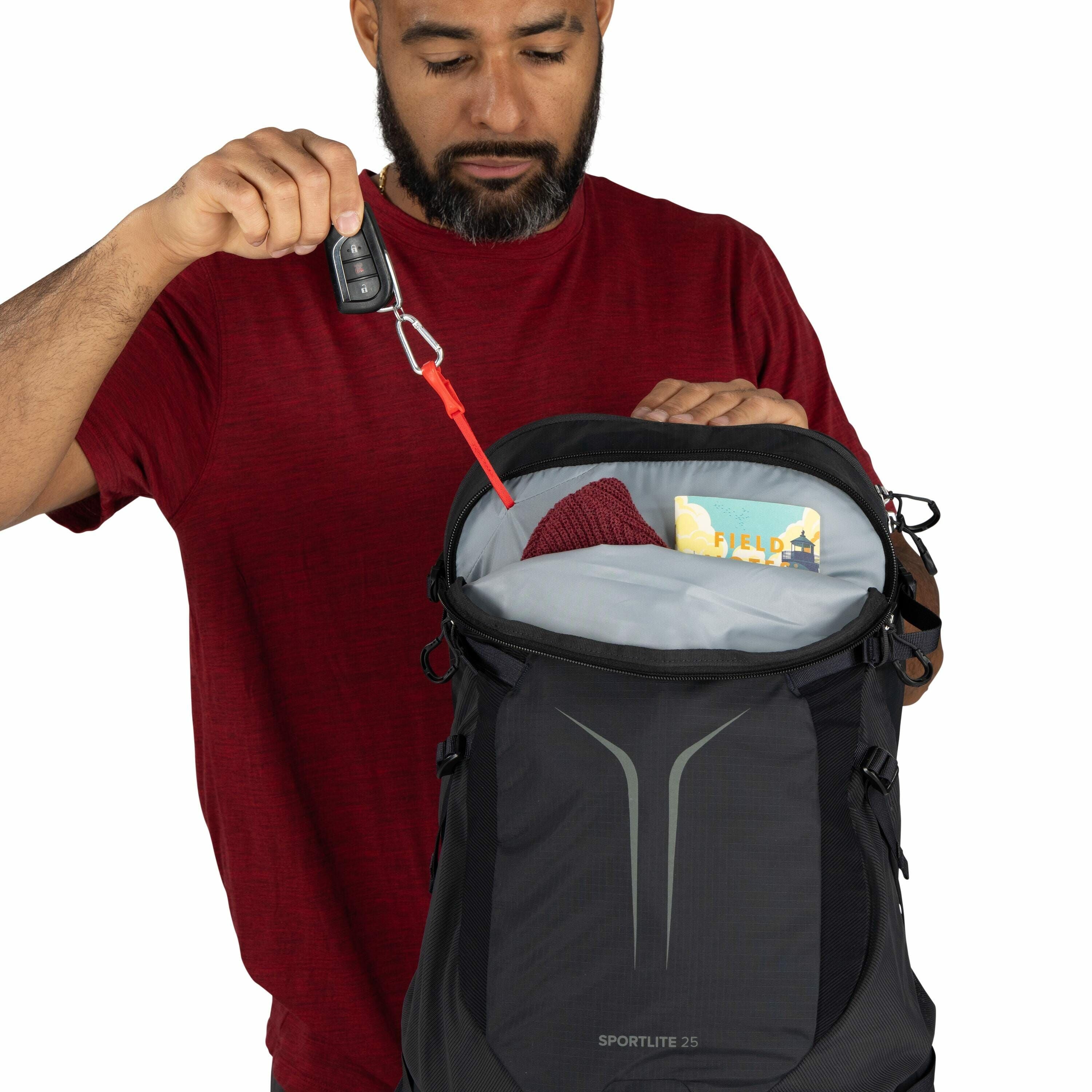 The image features a person wearing a red shirt, holding open a black backpack with "SPORTLITE 25" branding on it. The backpack is partially open, revealing its contents, which include a red knit item, likely a hat or scarf, and a yellow and blue booklet