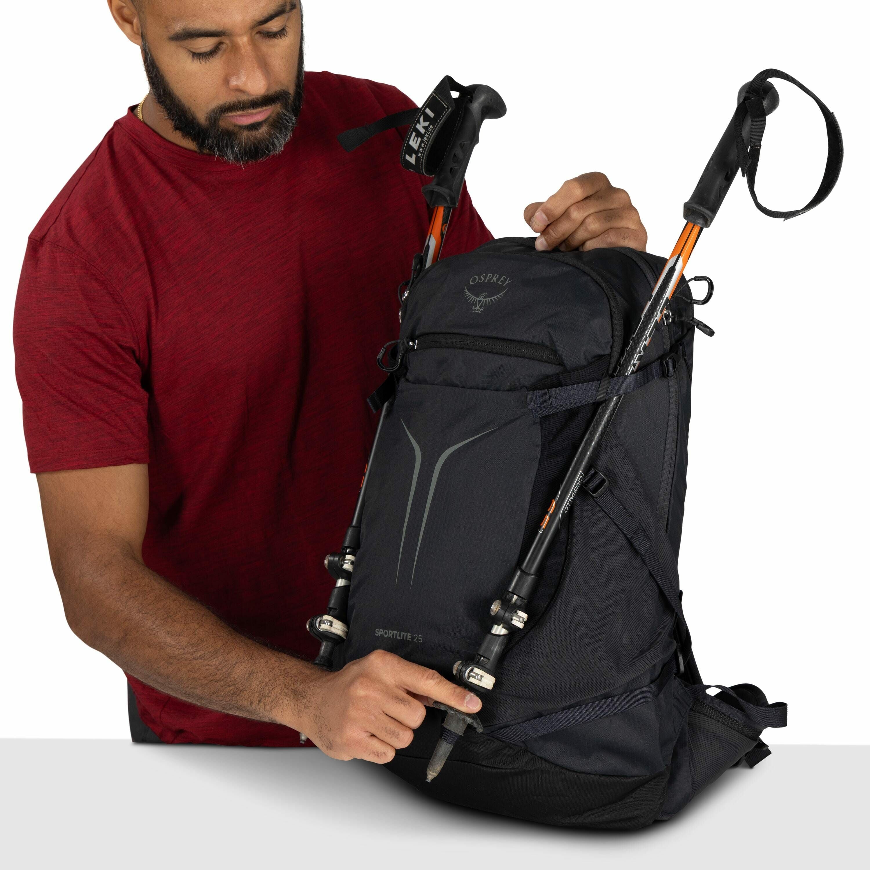The image shows a person wearing a red short-sleeved shirt holding a black Osprey backpack labeled "Sportlite 25." Attached to the backpack are two trekking poles, with one visible brand marking as "LEKI." The backpack features a sleek design with reflect