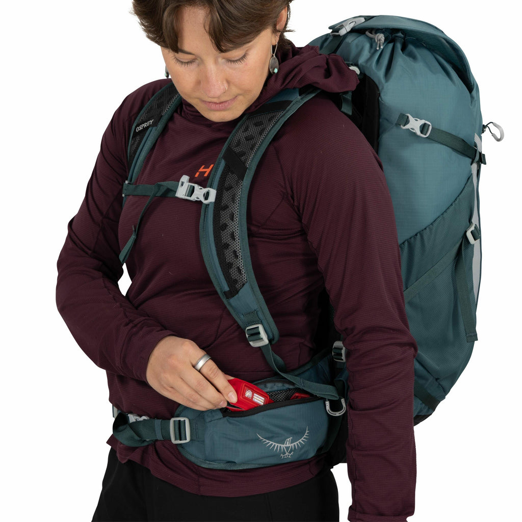 The image shows a person wearing a maroon long-sleeved top and black pants, equipped with an Osprey backpack. The backpack is teal in color and features adjustable straps for comfort and security. The person is reaching into the hip belt pocket of the bac