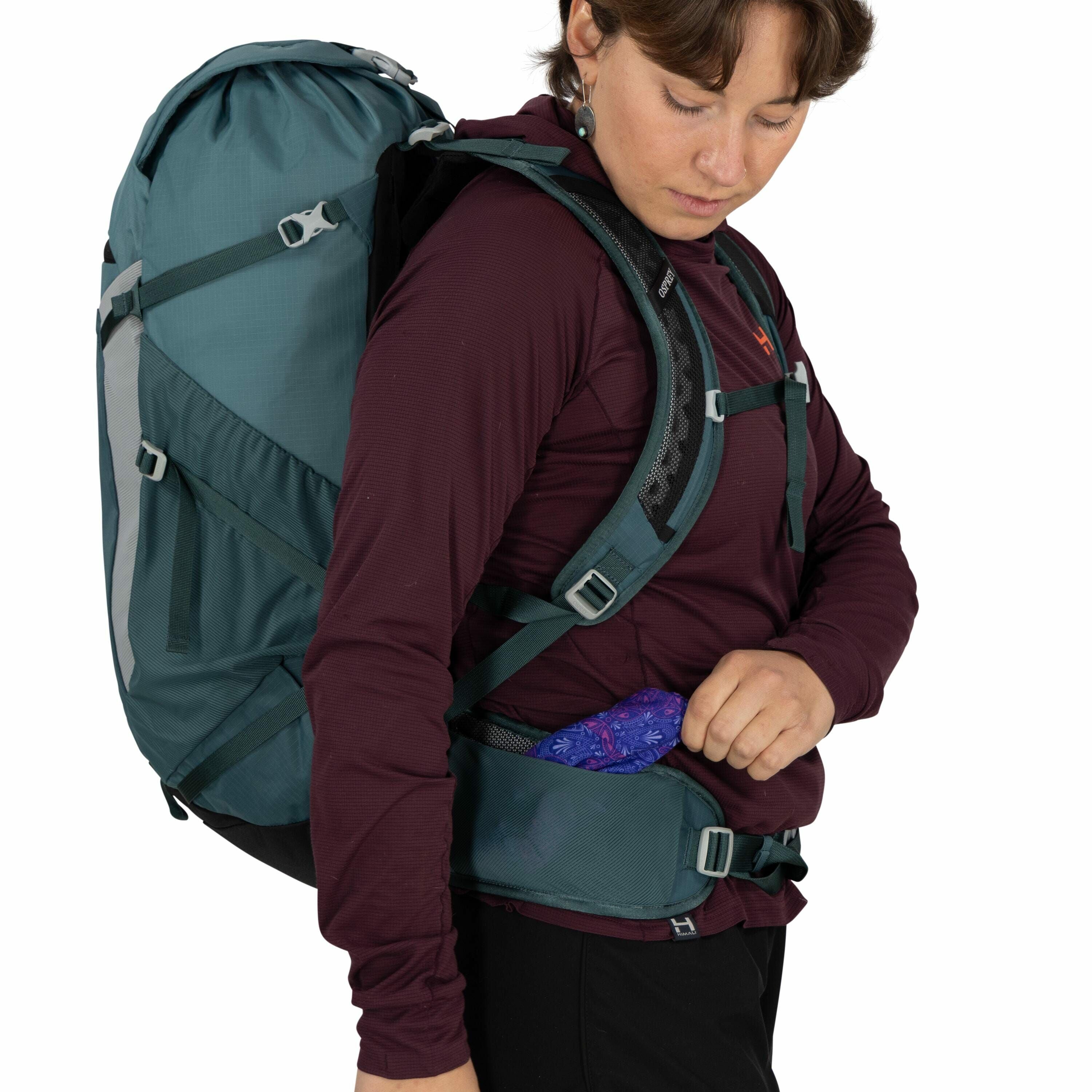 The image shows a person dressed in outdoor gear, wearing a dark maroon long-sleeve shirt and black pants. They are carrying a large, teal backpack equipped with padded shoulder straps and a hip belt, indicating readiness for a hike or camping trip. The b