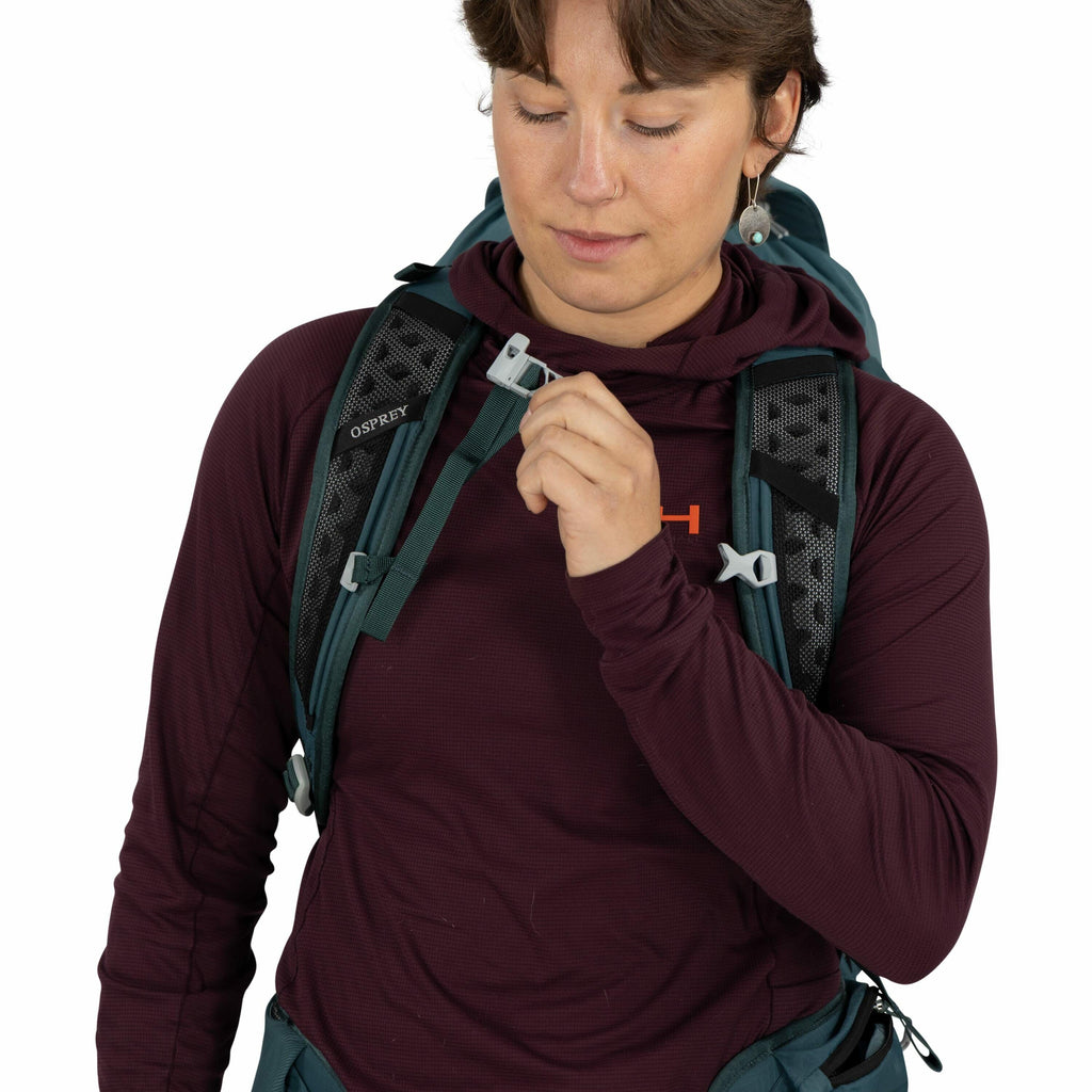 The image shows a person wearing a maroon long-sleeve hooded shirt with an orange "H" logo on it. They are wearing a dark teal backpack with the brand "Osprey" visible on the strap, featuring two gray plastic clips and mesh padding. The person is holding