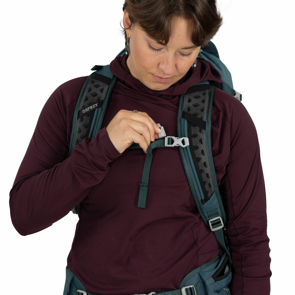 The image shows a person wearing a dark burgundy long-sleeve shirt while adjusting the strap of a teal and grey backpack. The backpack has well-padded shoulder straps and appears to be designed for hiking or outdoor use. The brand "Osprey" is visible on t