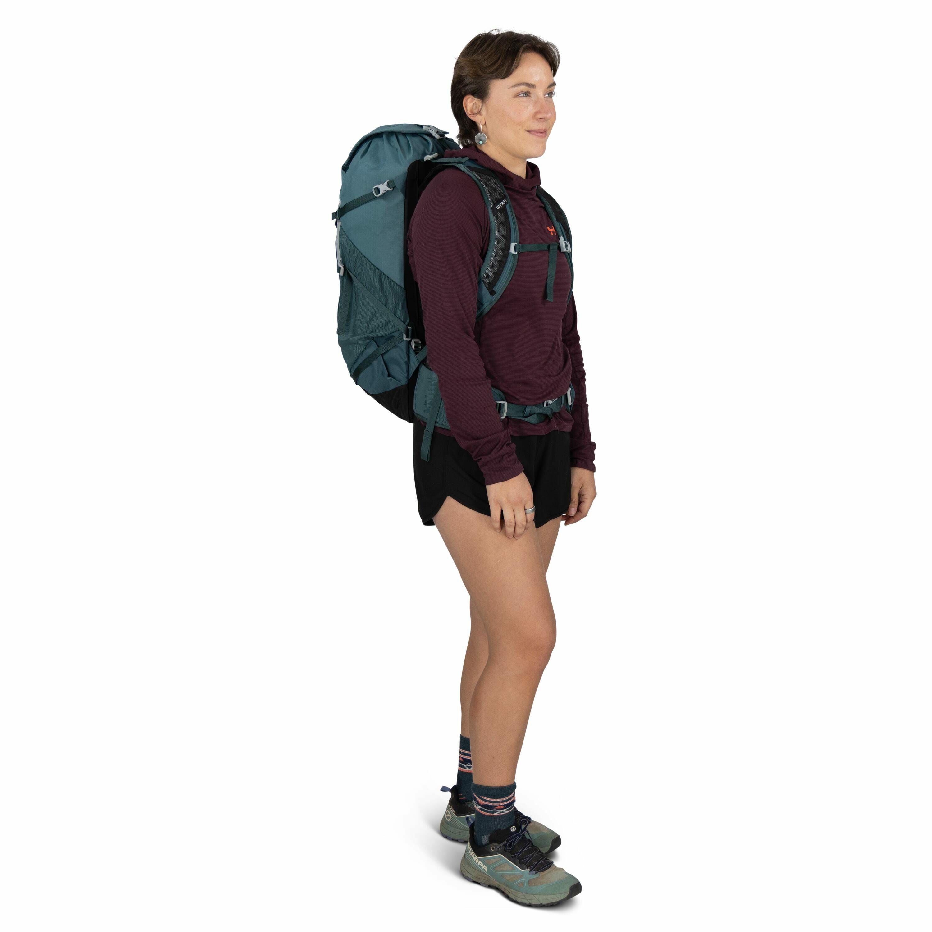 The image features a person standing in a side profile view, equipped for outdoor activities. They are wearing a maroon long-sleeve top and black shorts, with a large teal backpack strapped securely on their back. The backpack has multiple compartments an