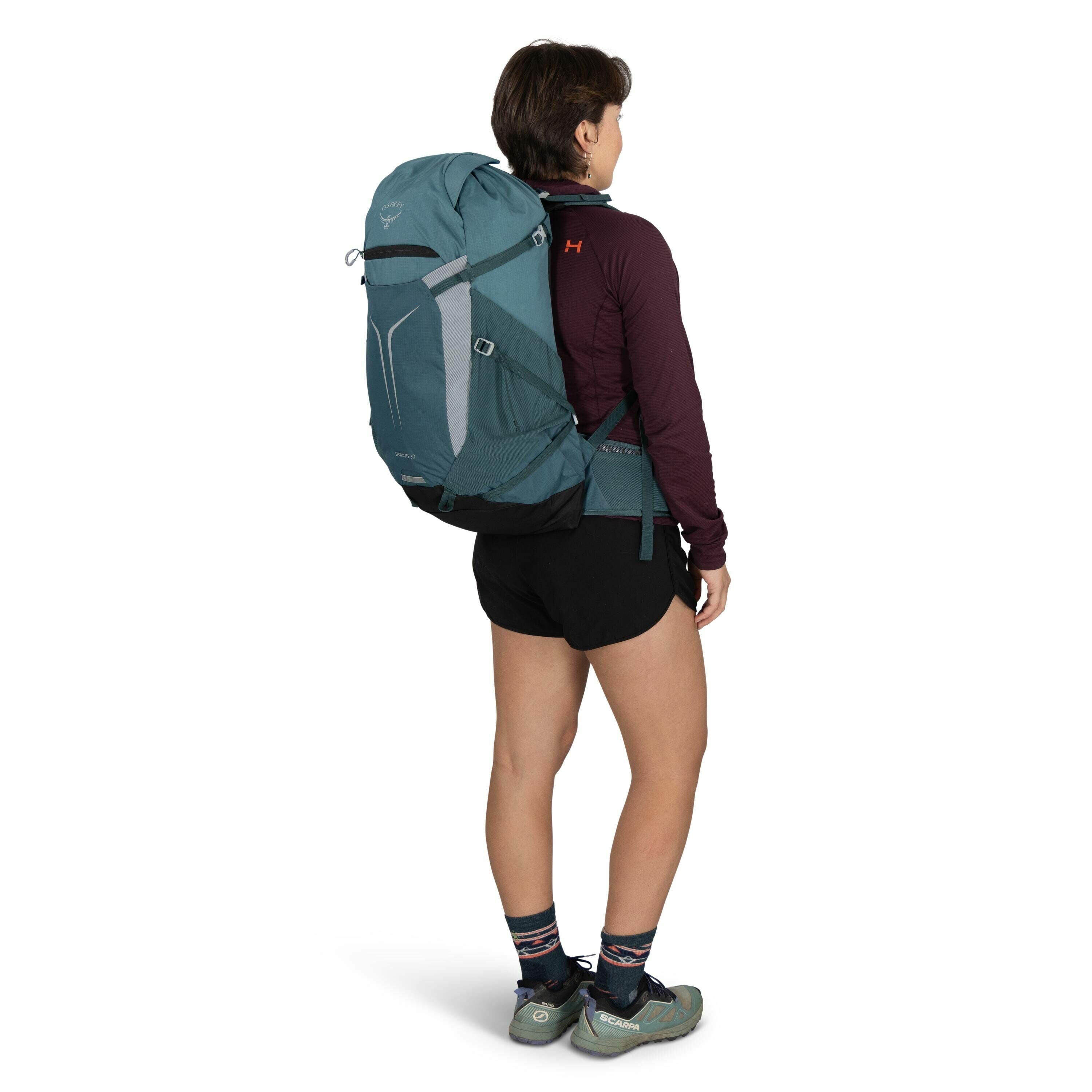 The image shows a person from the side, wearing a teal Osprey backpack. The individual is dressed in a maroon long-sleeved top, black shorts, and colorful socks, paired with green Scarpa hiking shoes. The Osprey logo is visible on the top part of the back