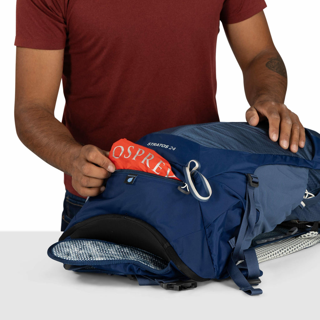 The image shows a man wearing a red shirt handling a blue Osprey backpack with a "STRATOS 24" label visible near the top. He is pulling out a red rain cover with the Osprey logo from a zippered compartment on the front of the backpack. The backpack appear