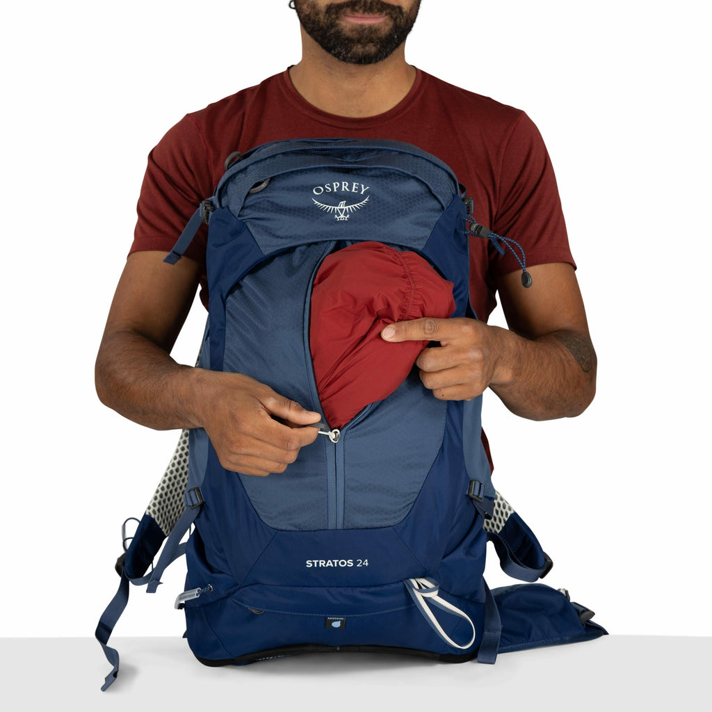 The image features a man wearing a red T-shirt demonstrating the functionality of an Osprey Stratos 24 backpack. He is partially unzipping a front compartment, revealing a red item inside, likely a built-in rain cover or an accessory pouch. The backpack i