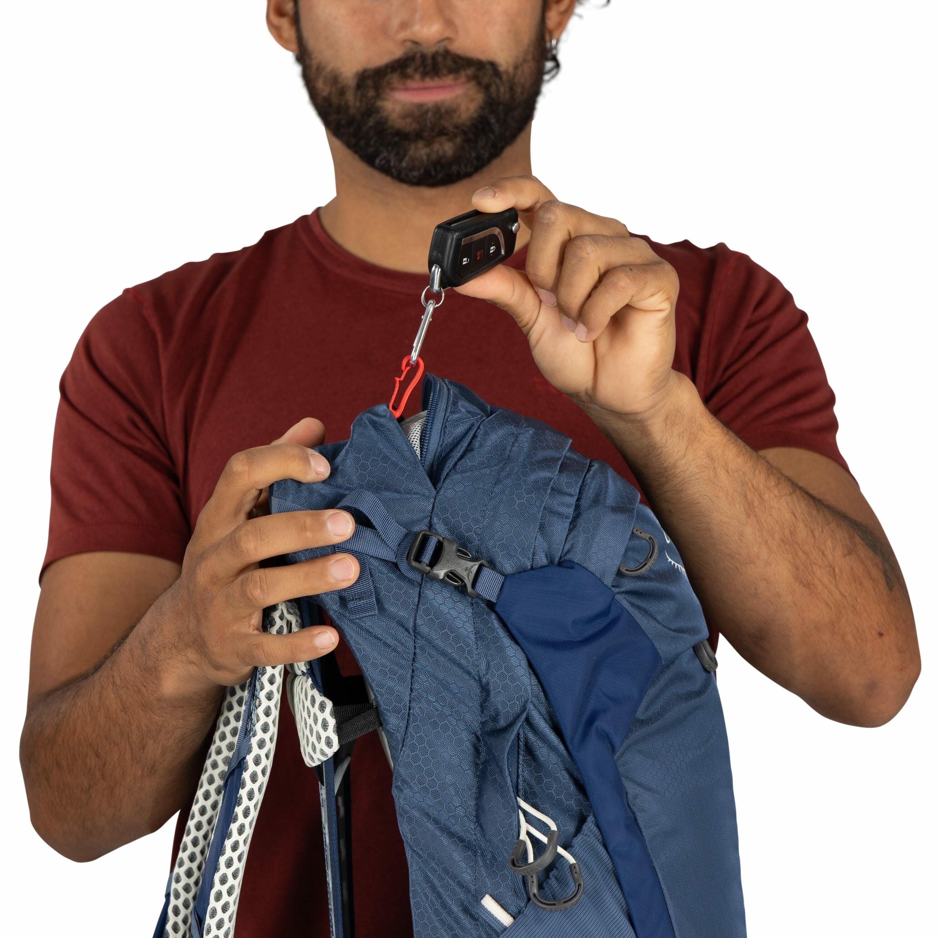 The image depicts a person wearing a red t-shirt, holding a dark blue backpack with a patterned design. The individual is using a handheld luggage scale to weigh the backpack. The scale is black and has a small digital display with buttons. A red hook is