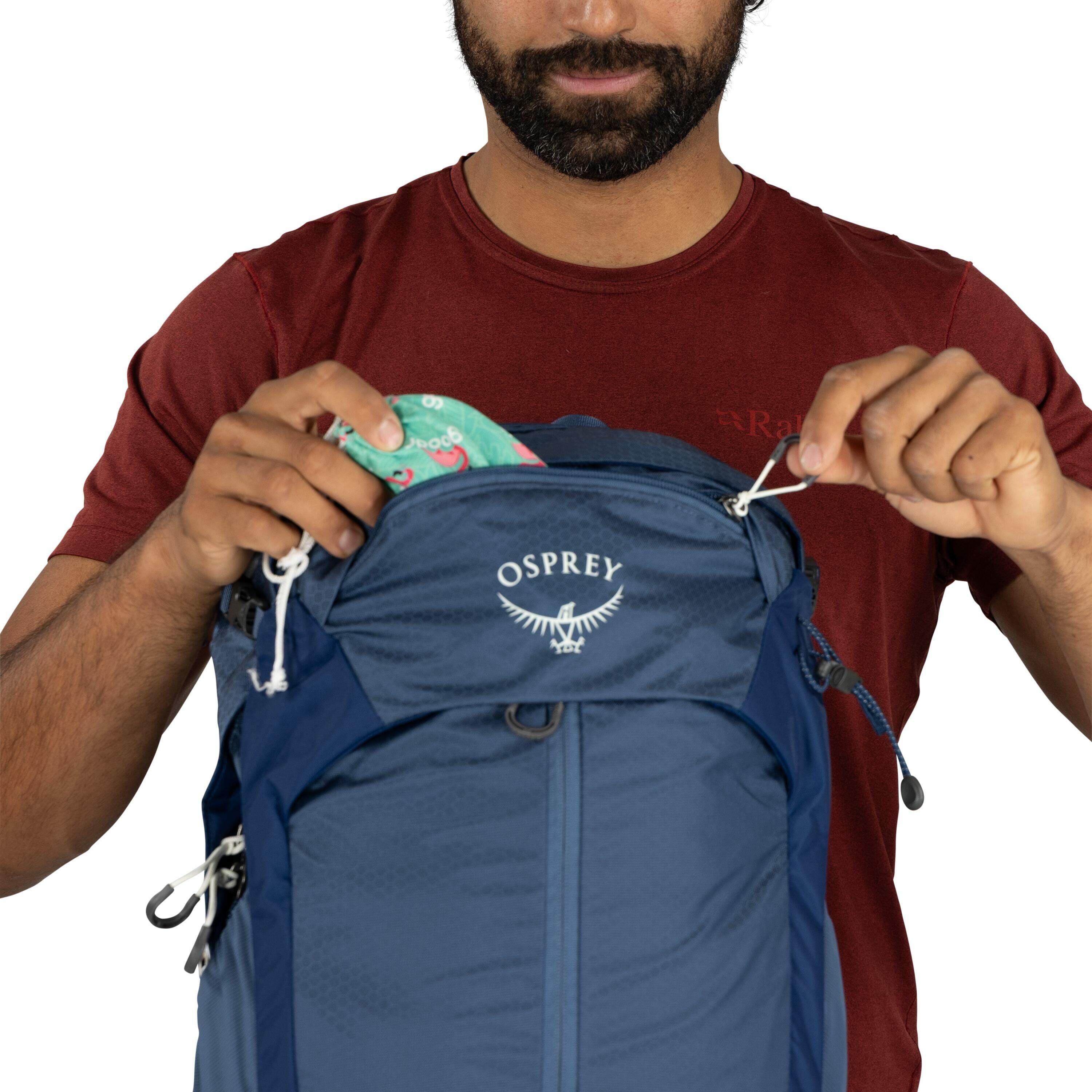 The image features a person wearing a reddish-brown t-shirt, partially visible from the shoulders up, as they interact with a blue Osprey backpack. The person is holding the zippers of the backpack's top compartment, which is open, revealing a colorful fa