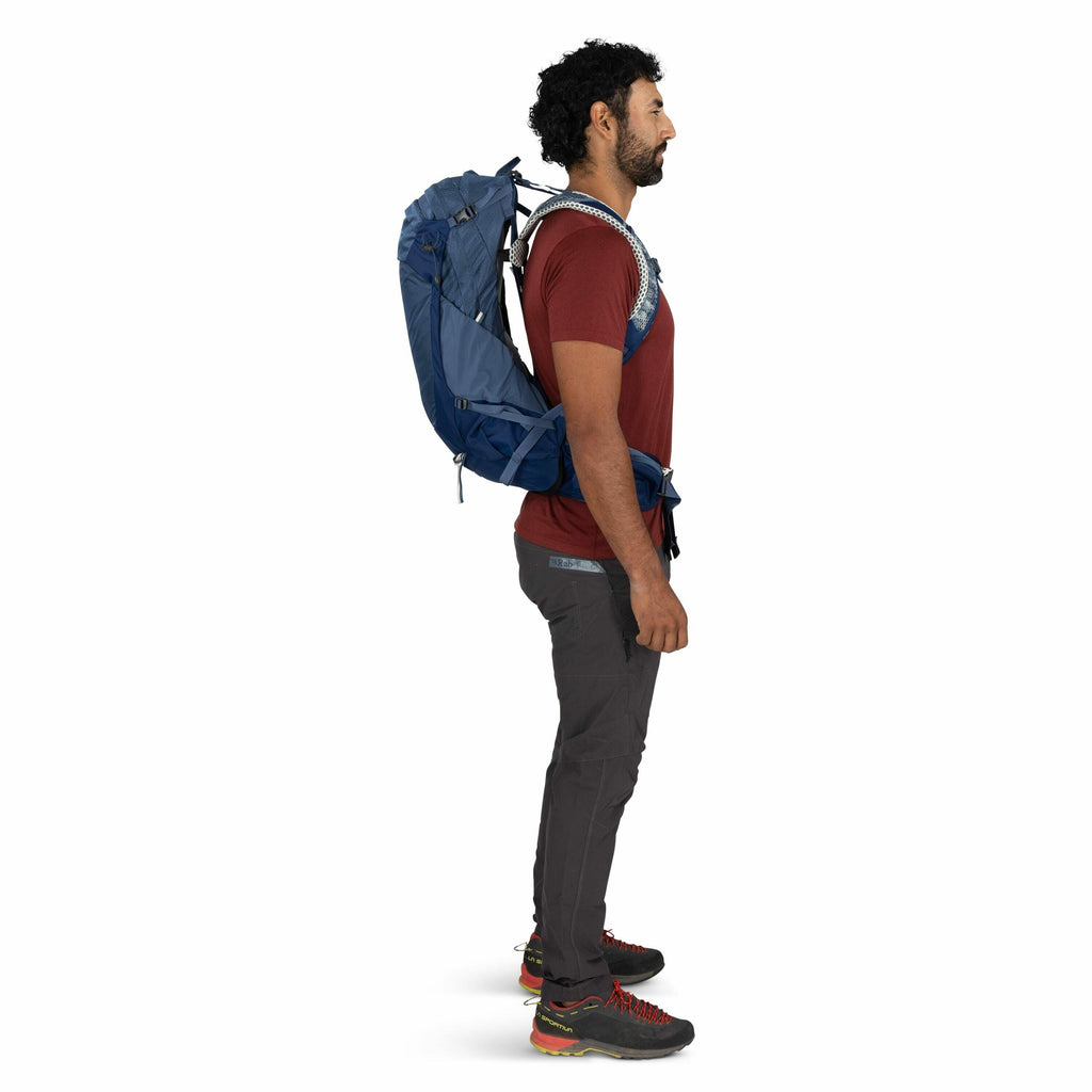 The image features a person standing in a side profile view, wearing a hiking outfit. The individual is dressed in a maroon short-sleeve shirt, gray hiking pants, and trail shoes with black, red, and yellow accents. They are carrying a blue backpack, whic