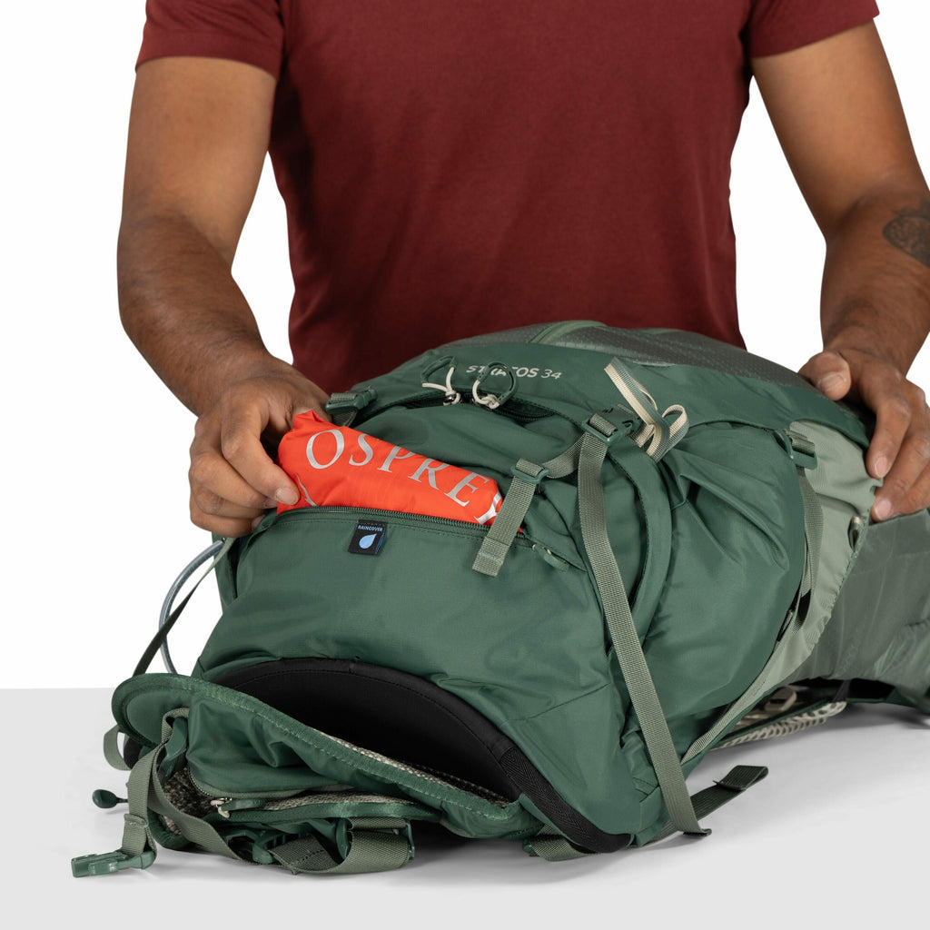 The image shows a person wearing a maroon short-sleeve shirt, demonstrating the use of a green Osprey backpack. The individual is pulling out an orange item, likely a rain cover, from a front compartment labeled with the Osprey logo. The backpack is resti