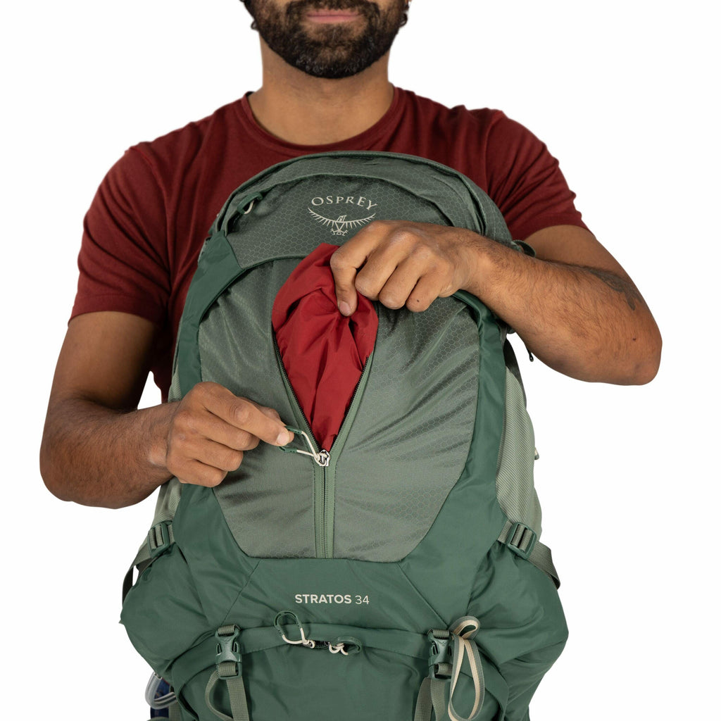 The image shows a man wearing a red t-shirt and holding a green Osprey backpack. The backpack is labeled "STRATOS 34" and is partially unzipped, revealing a red fabric inside. The man is pulling the zipper down, using one hand to hold the zipper pull and