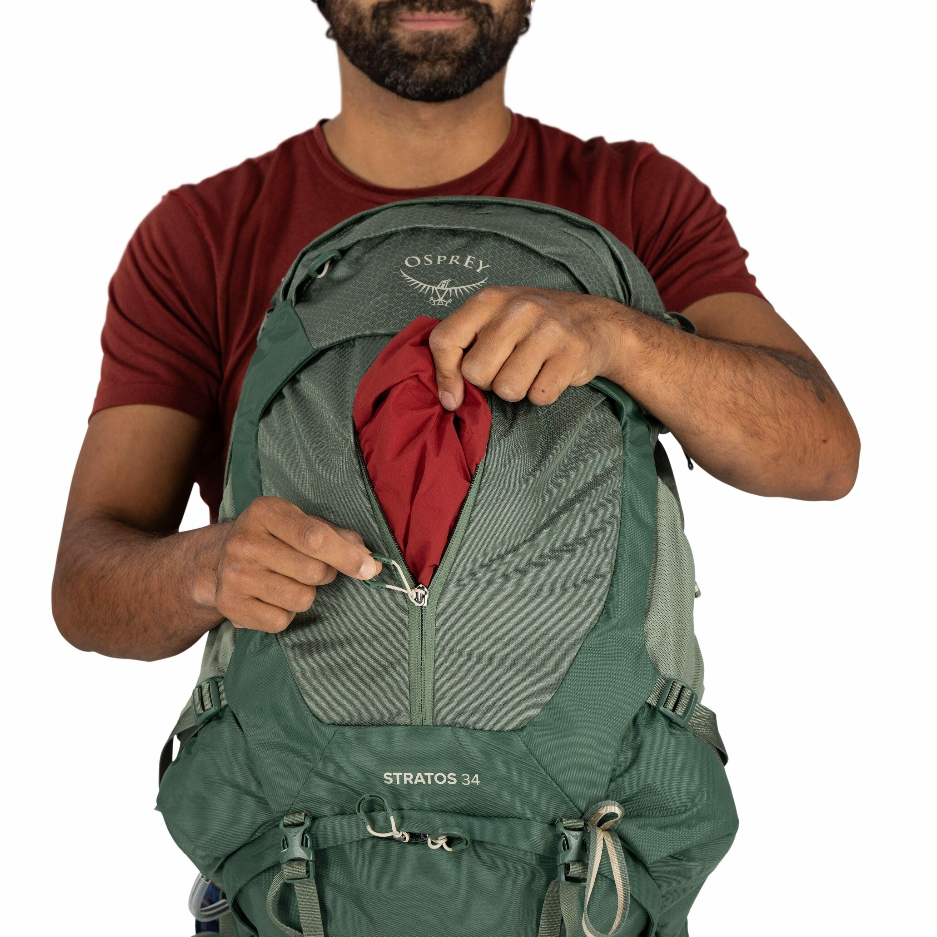 The image shows a man wearing a red t-shirt and holding a green Osprey backpack. The backpack is labeled "STRATOS 34" and is partially unzipped, revealing a red fabric inside. The man is pulling the zipper down, using one hand to hold the zipper pull and