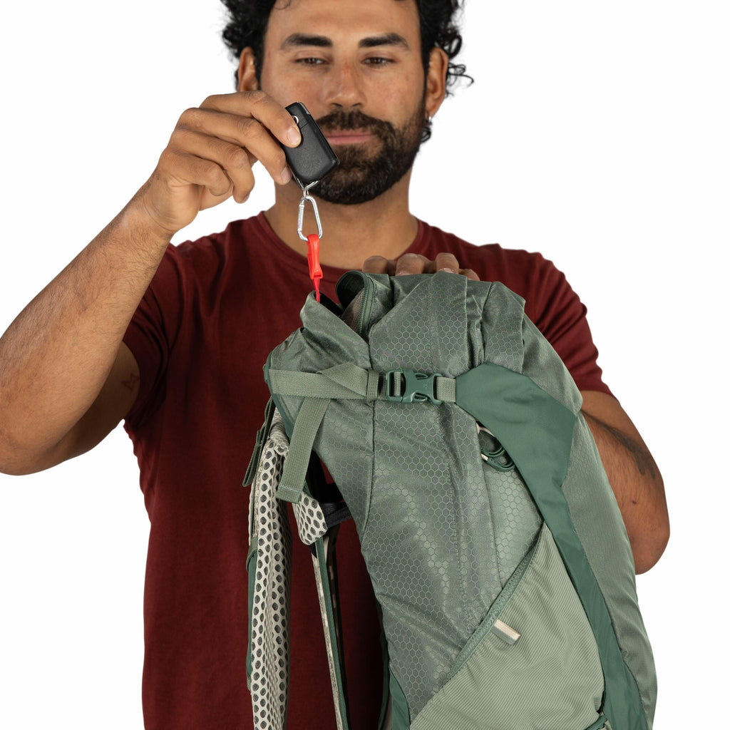 The image depicts a person holding a green backpack against a plain white background. The person is wearing a dark red short-sleeved shirt and has a small tattoo on their right arm. They are holding the top of the backpack with one hand, while the other h