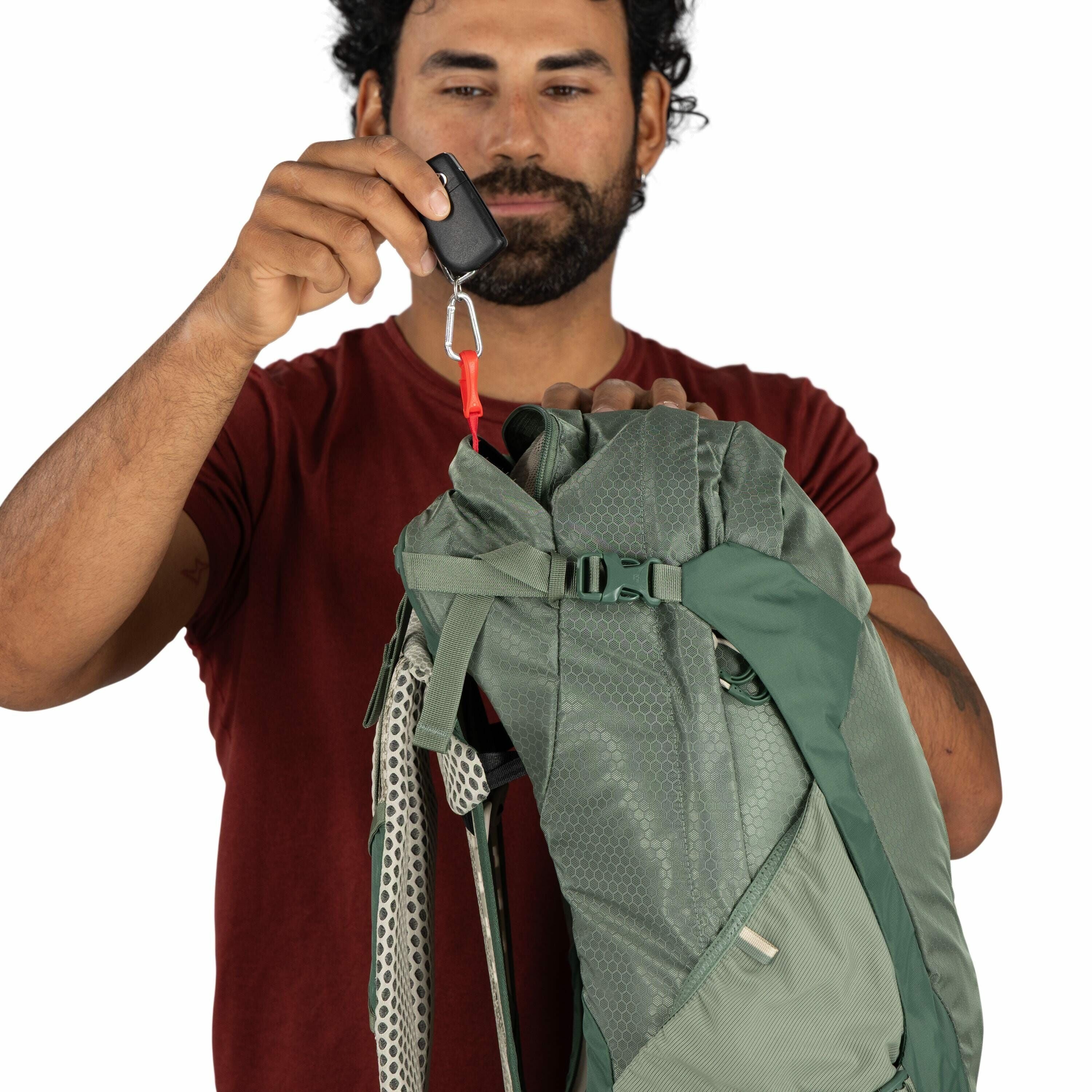 The image depicts a person holding a green backpack against a plain white background. The person is wearing a dark red short-sleeved shirt and has a small tattoo on their right arm. They are holding the top of the backpack with one hand, while the other h