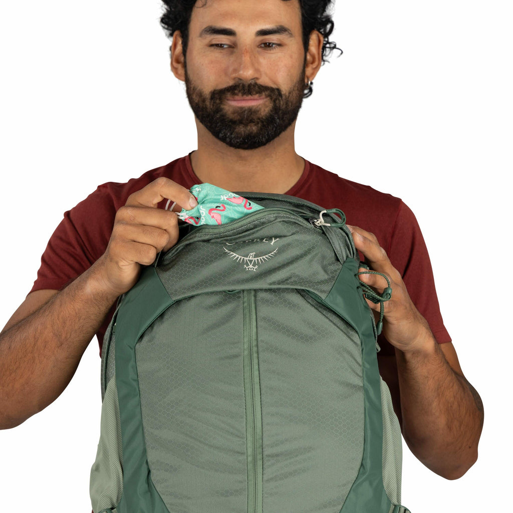 The image shows a person wearing a red shirt holding a green Osprey backpack. The individual is in the process of placing or retrieving a colorful item with a teal and pink flamingo pattern from the backpack's top pocket. The backpack features the Osprey