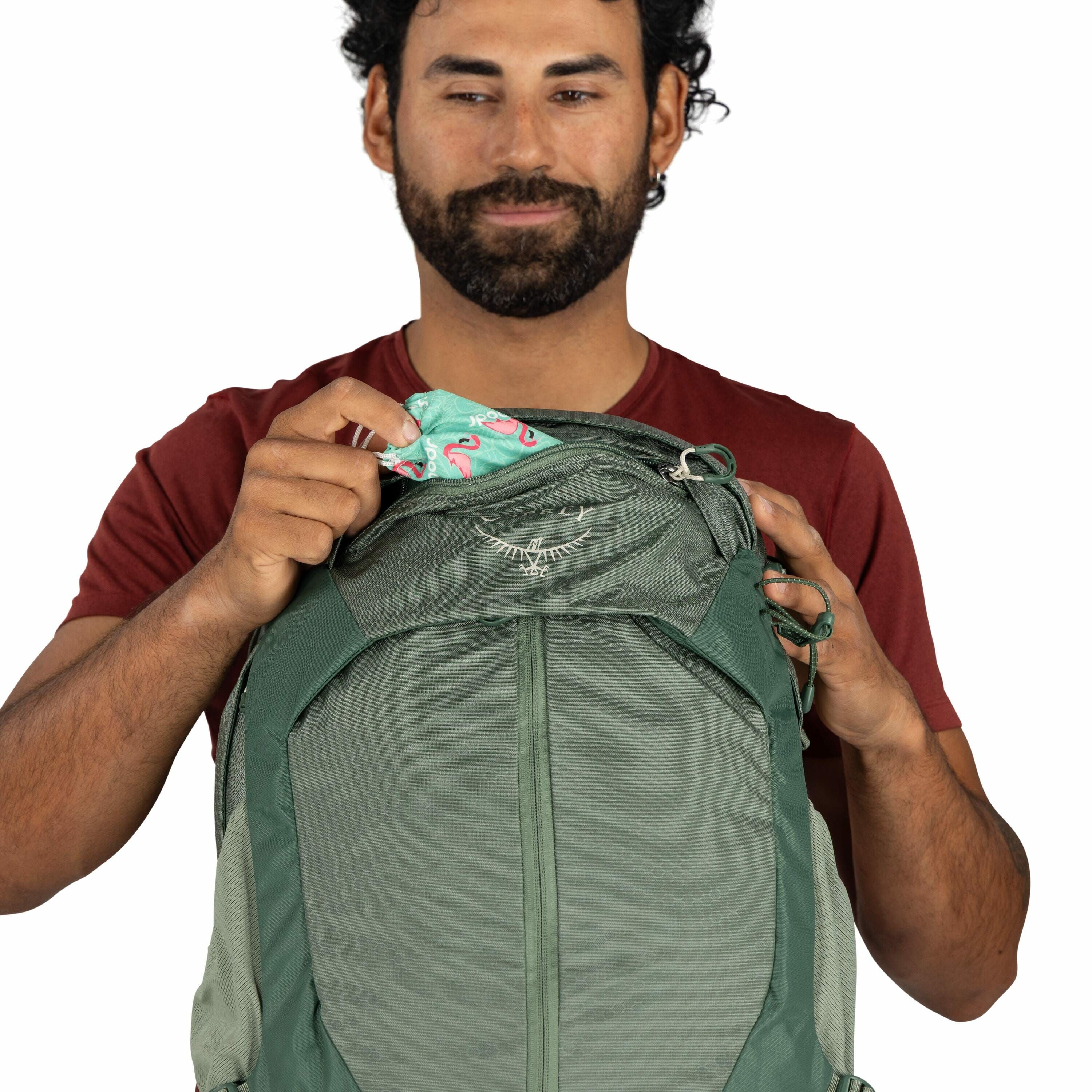 The image shows a person wearing a red shirt holding a green Osprey backpack. The individual is in the process of placing or retrieving a colorful item with a teal and pink flamingo pattern from the backpack's top pocket. The backpack features the Osprey