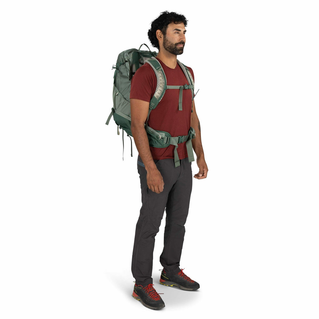 The image features a person dressed in outdoor attire, likely prepared for hiking or trekking. They are wearing a red short-sleeve t-shirt and dark gray hiking pants. On their feet are black shoes with red laces and subtle green accents, suggesting they a