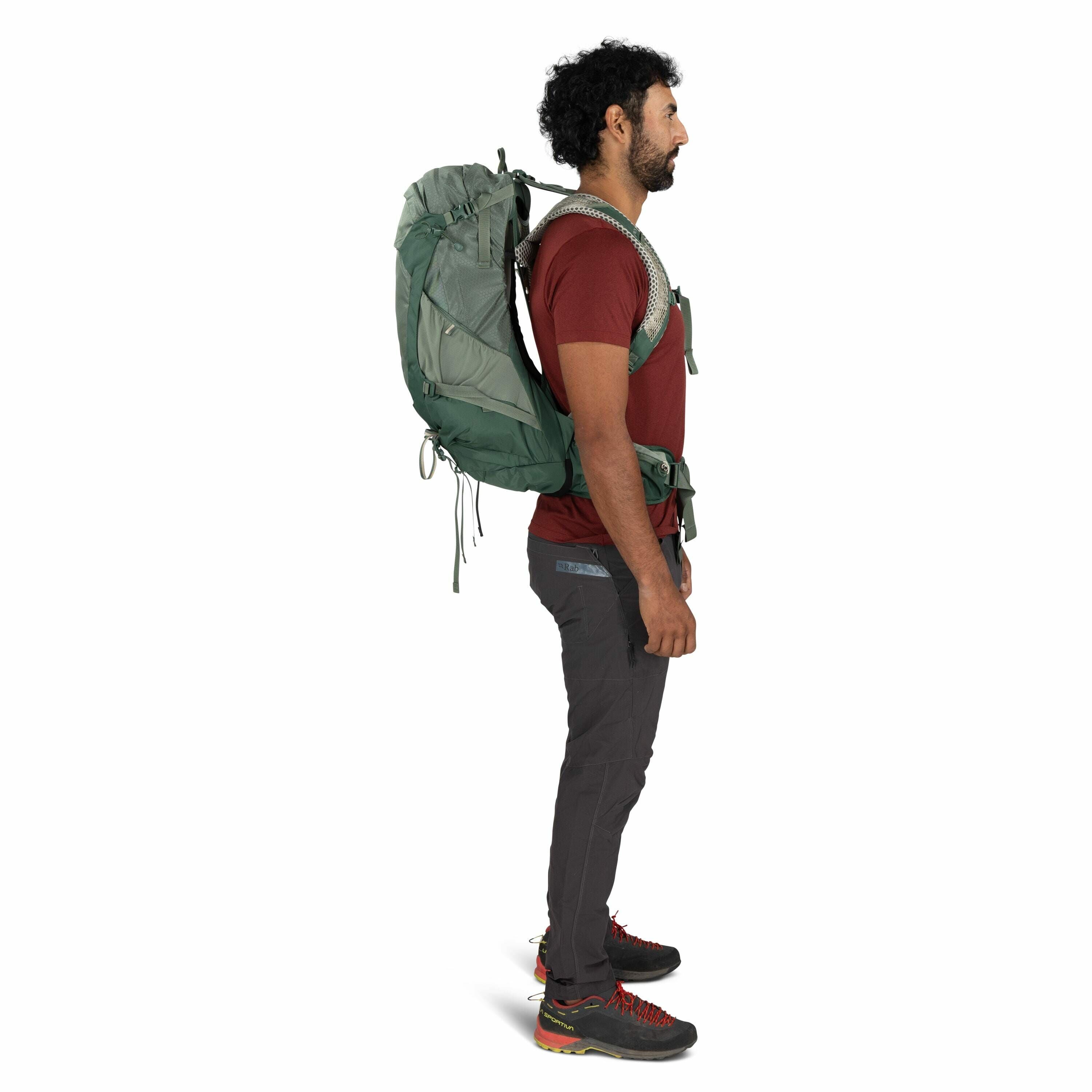 The image shows a man standing in profile wearing hiking gear, including a large green backpack, a red t-shirt, dark gray pants, and hiking boots. The backpack appears to be well-fitted with adjustable straps and a waist belt for support. His pants have a