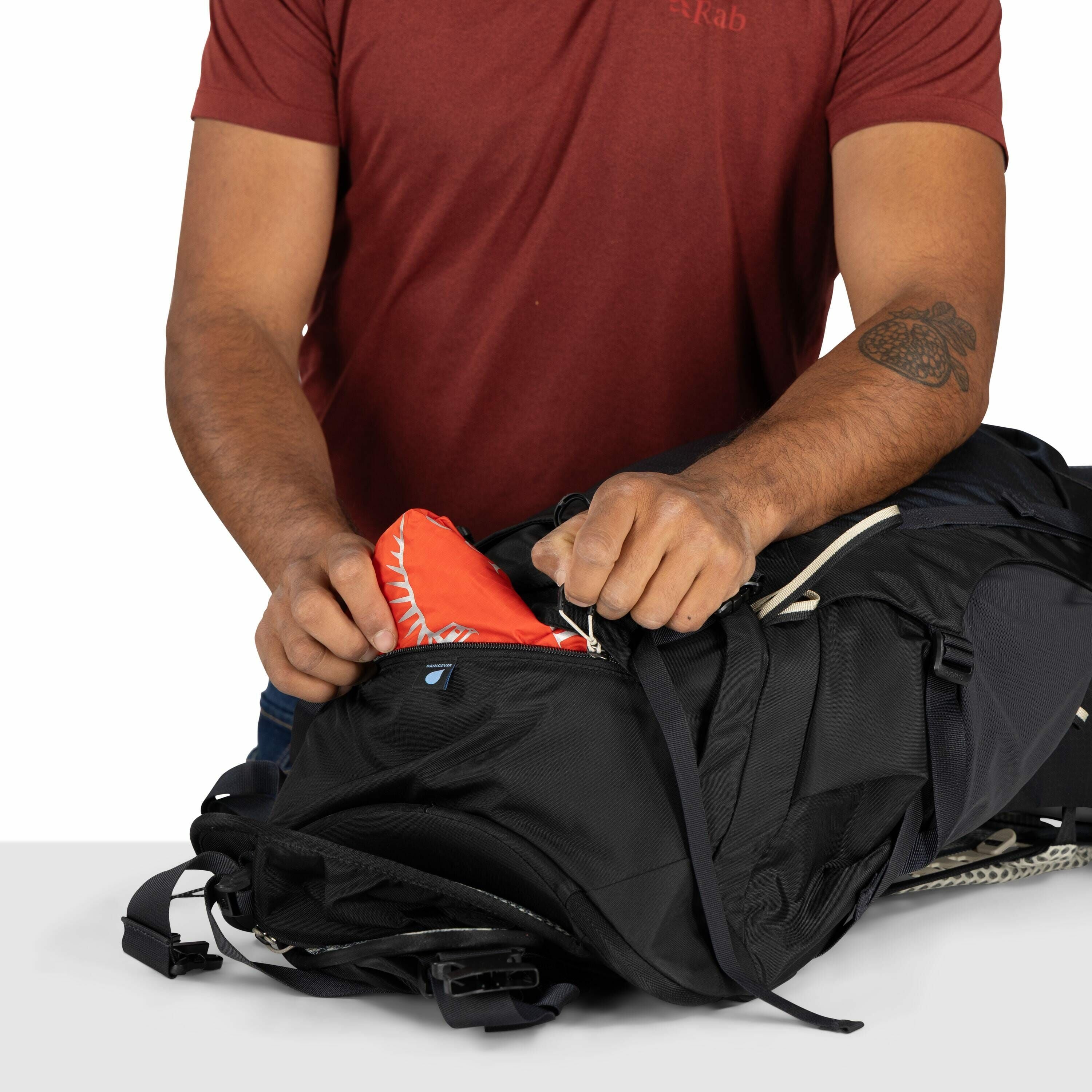 The image shows a person wearing a red Rab-branded T-shirt while reaching into a black backpack. The person has a tattoo on their left forearm, and they are pulling out a red item from one of the backpack's compartments. The backpack appears to be designe