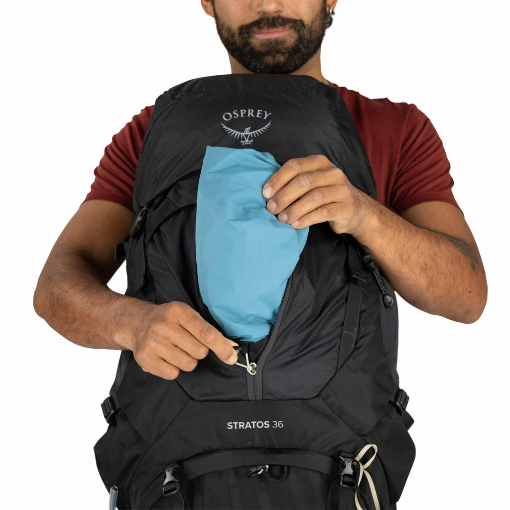 The image features a man with a beard and wearing a red shirt, holding a black Osprey backpack. He is using one hand to insert or retrieve a light blue item from a zippered compartment on the front of the backpack. The Osprey logo, featuring an abstract b