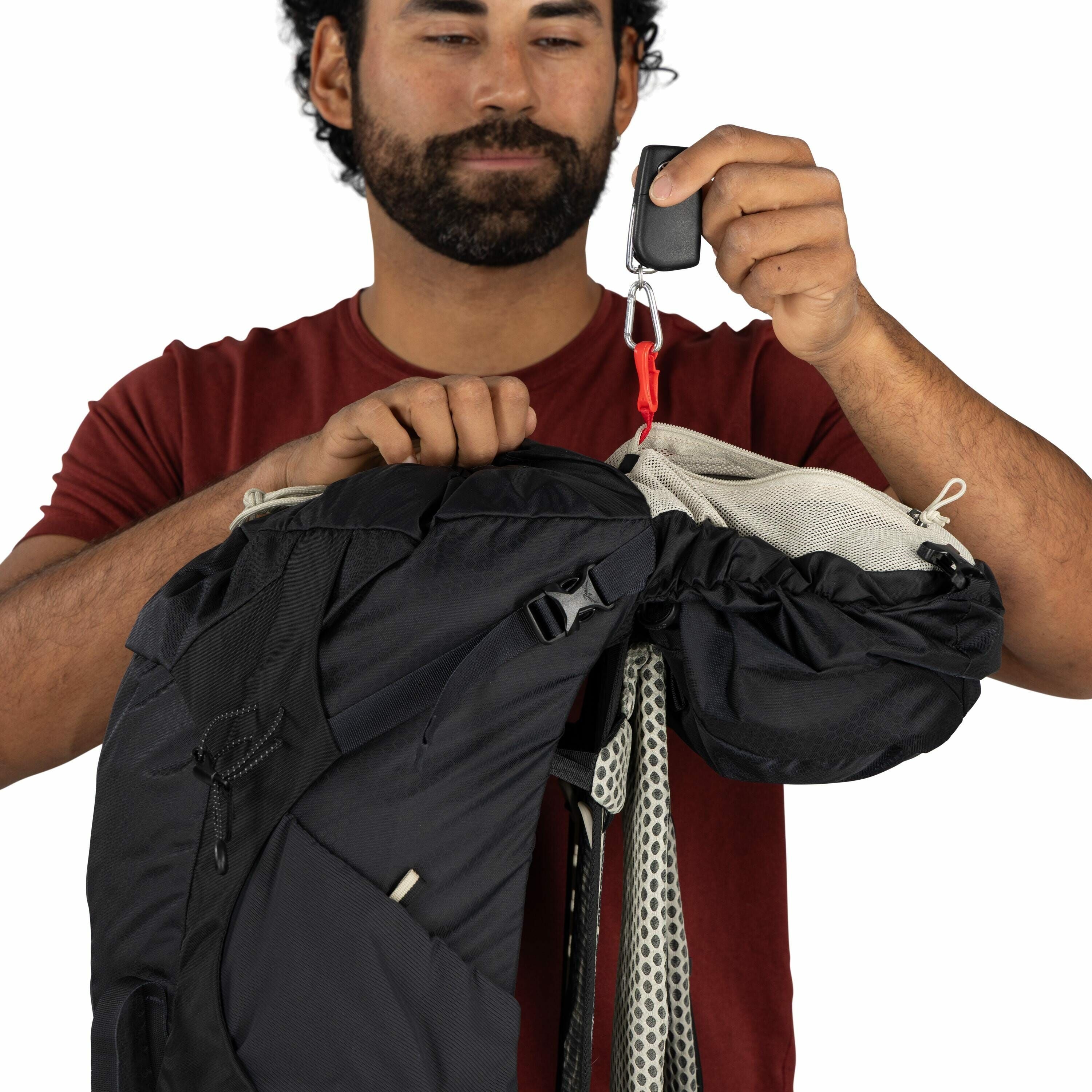 The image shows a man wearing a red shirt holding a black backpack in one hand. The backpack is open, revealing an interior mesh pocket with a drawstring closure. The man is holding a small black device with a carabiner clip attached to a red strap connec