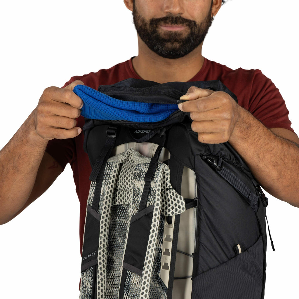 The image shows a person holding a backpack in front of them with both hands. The person is wearing a red T-shirt, and part of their hands and arms are visible. The backpack is predominantly black, and the brand "Osprey" is visible on the front. The bag's