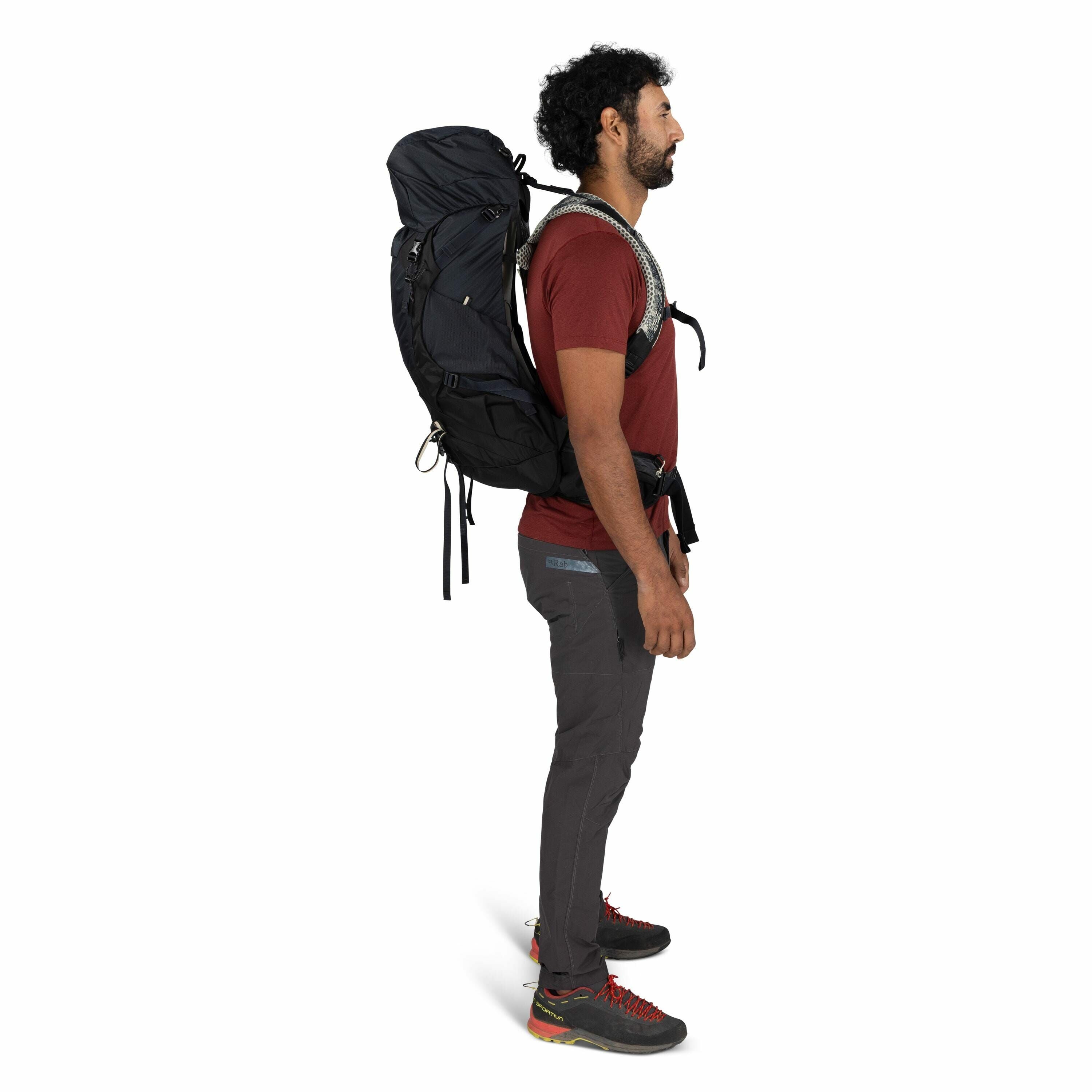 The image shows a man standing in profile, carrying a large hiking backpack. He is dressed in outdoor apparel, including a red short-sleeved t-shirt and dark gray hiking pants, which feature a logo from "Arc'teryx". The man is also wearing black hiking sh