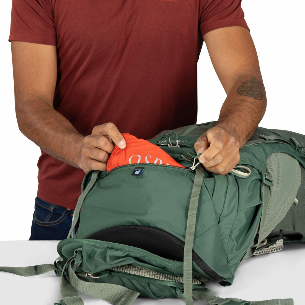 The image shows a person with medium-toned skin wearing a maroon T-shirt, who is packing or unpacking an orange item from a green backpack. The orange item is partially visible and has the letters "OSP" on it, suggesting it could be an Osprey-branded item