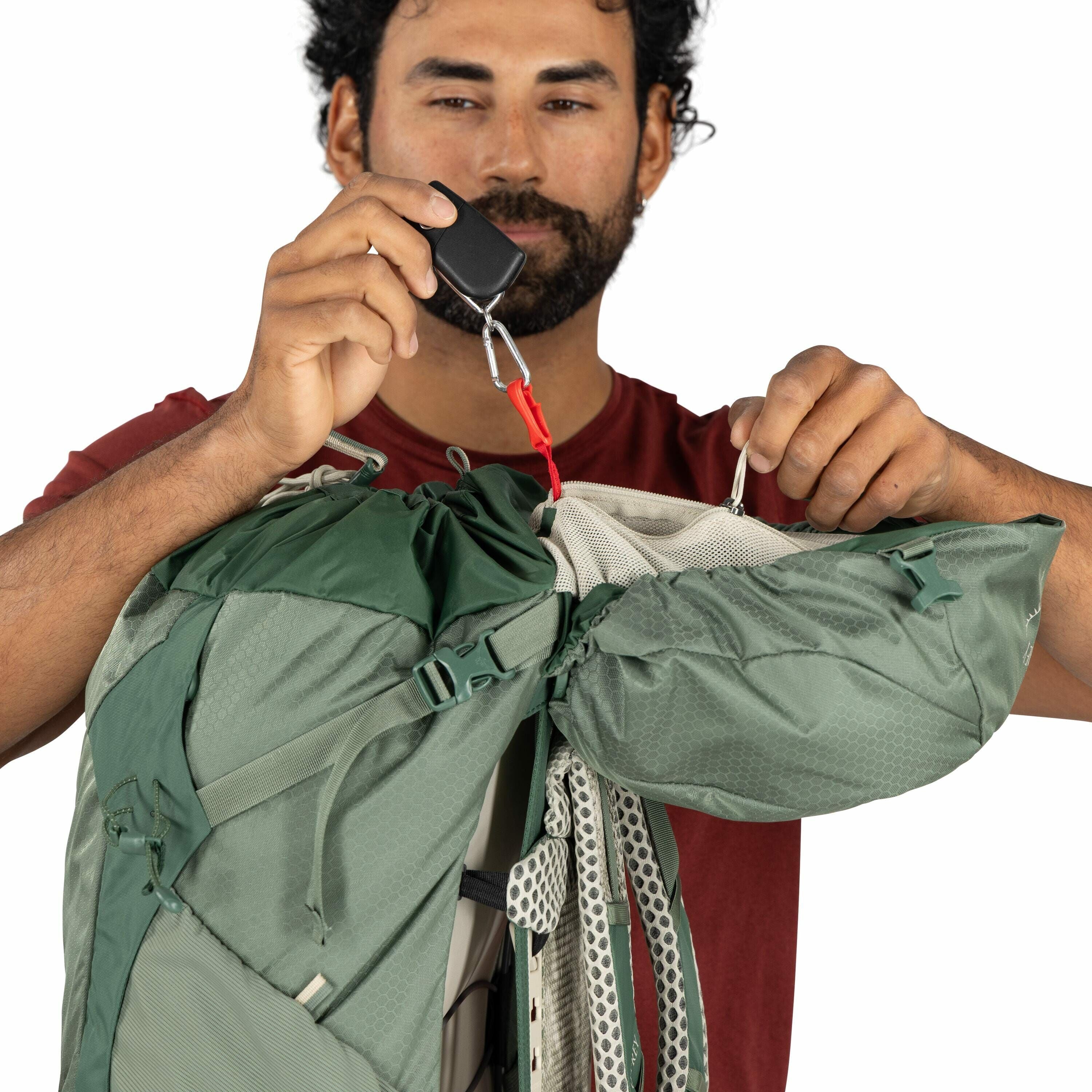 The image shows a person holding a green backpack, using two carabiners to attach or secure parts of the bag. The individual is wearing a red shirt, and their hands are seen adjusting the carabiners and the backpack's straps. The backpack is a large, outd
