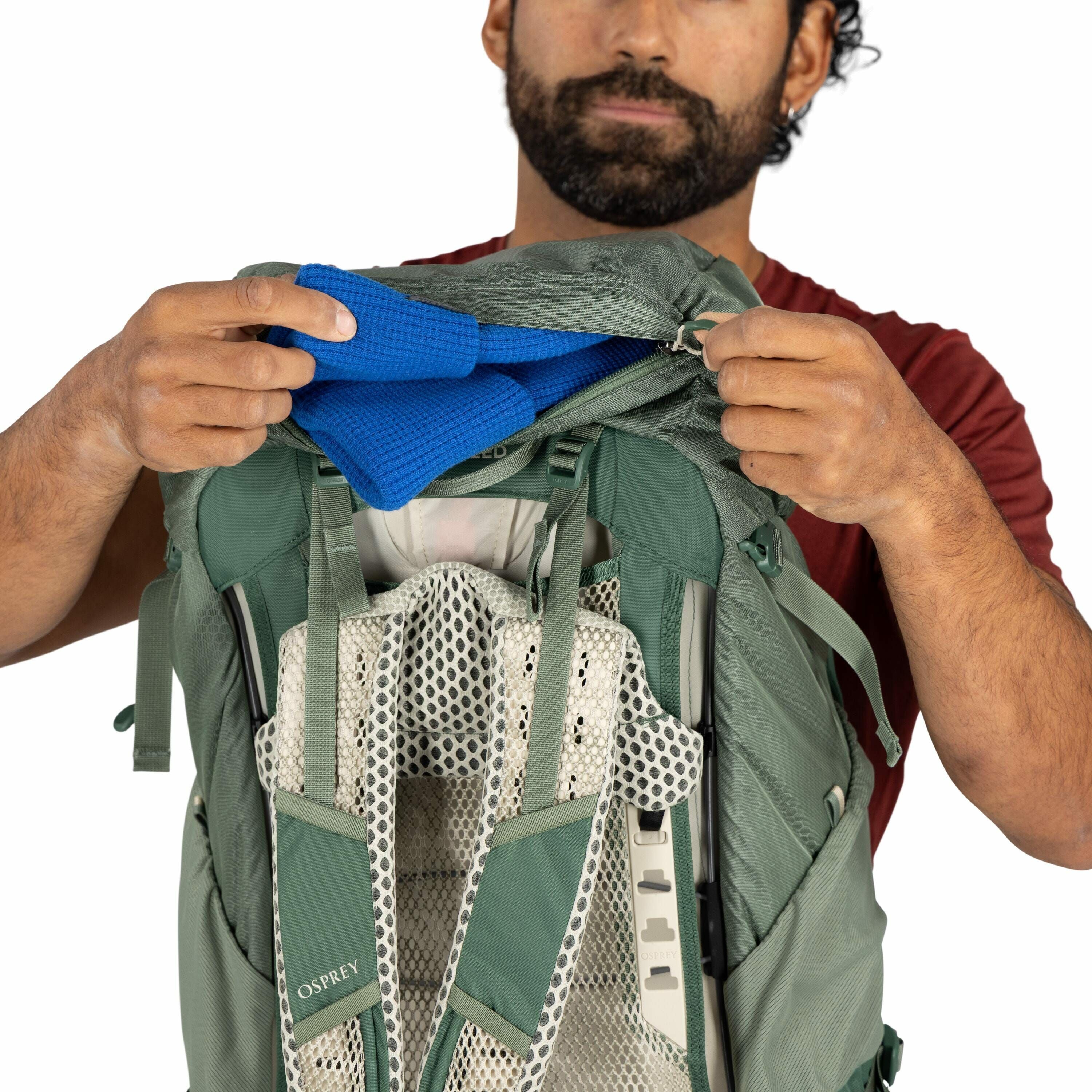 The image shows a person holding a green backpack with a mesh and padded harness system designed for comfort and ventilation. The backpack brand is "Osprey," visible on the shoulder strap. The individual is unzipping the main compartment of the backpack a