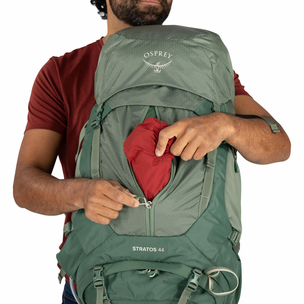The image shows a person wearing a red short-sleeve shirt and holding a green backpack from the Osprey brand. The backpack has the model name "Stratos 44" written on it, and the person is unzipping a front pocket while placing a red item, possibly a jacke