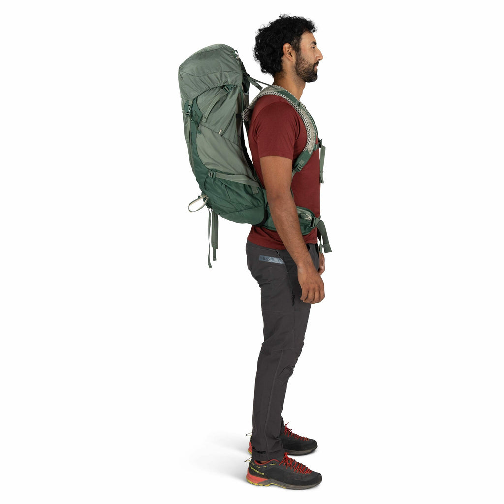 The image features a man standing sideways, wearing a large green hiking backpack. He is dressed in outdoor gear, including a red short-sleeve shirt, dark gray pants, and black hiking shoes with red accents. The backpack appears sturdy and is strapped sec