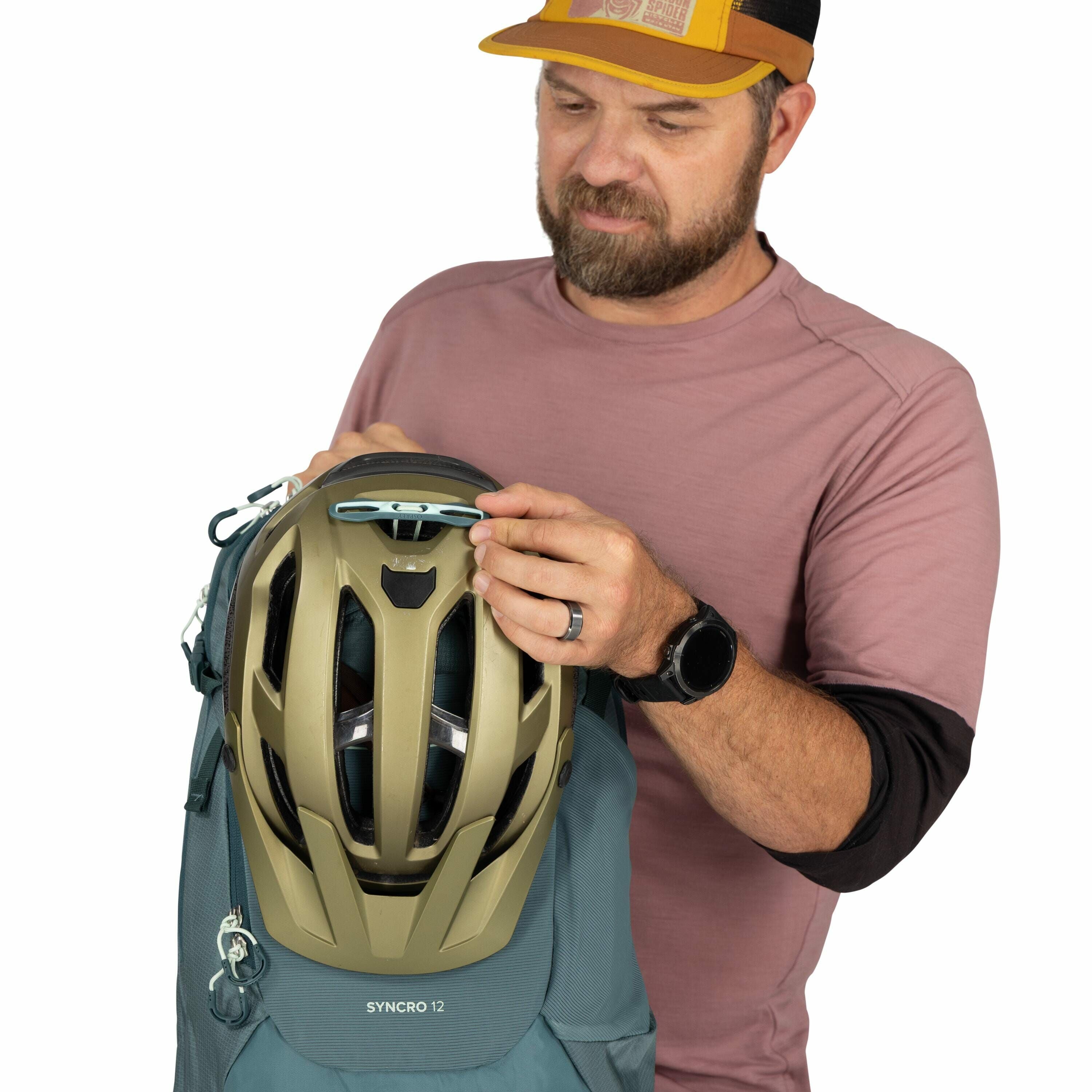 In the image, a person is holding a gray backpack with a helmet attached to the front using a blue clip. The backpack is labeled "SYNCRO 12" and has several buckles and straps. The helmet is a gold color with a ventilation design on its surface. The perso