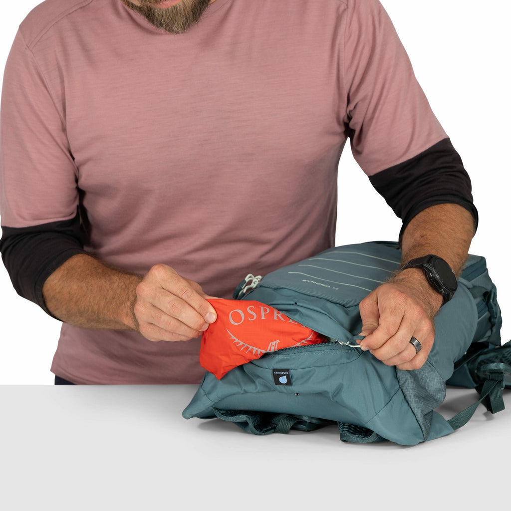 The image shows a person wearing a pink long-sleeve shirt with black sleeves underneath, seated at a table. They are holding a light blue Osprey backpack and pulling out a bright orange pouch with the Osprey logo on it from an exterior pocket. The backpac