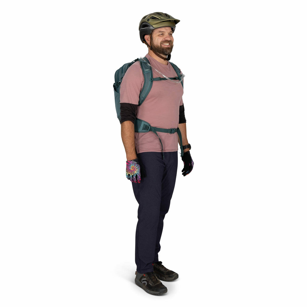 The image shows a person standing and dressed for outdoor or cycling activities. They are wearing a bicycle helmet, a light pink short-sleeve t-shirt over a long-sleeve black undershirt, and dark pants. They have a pair of colorful, patterned gloves on th