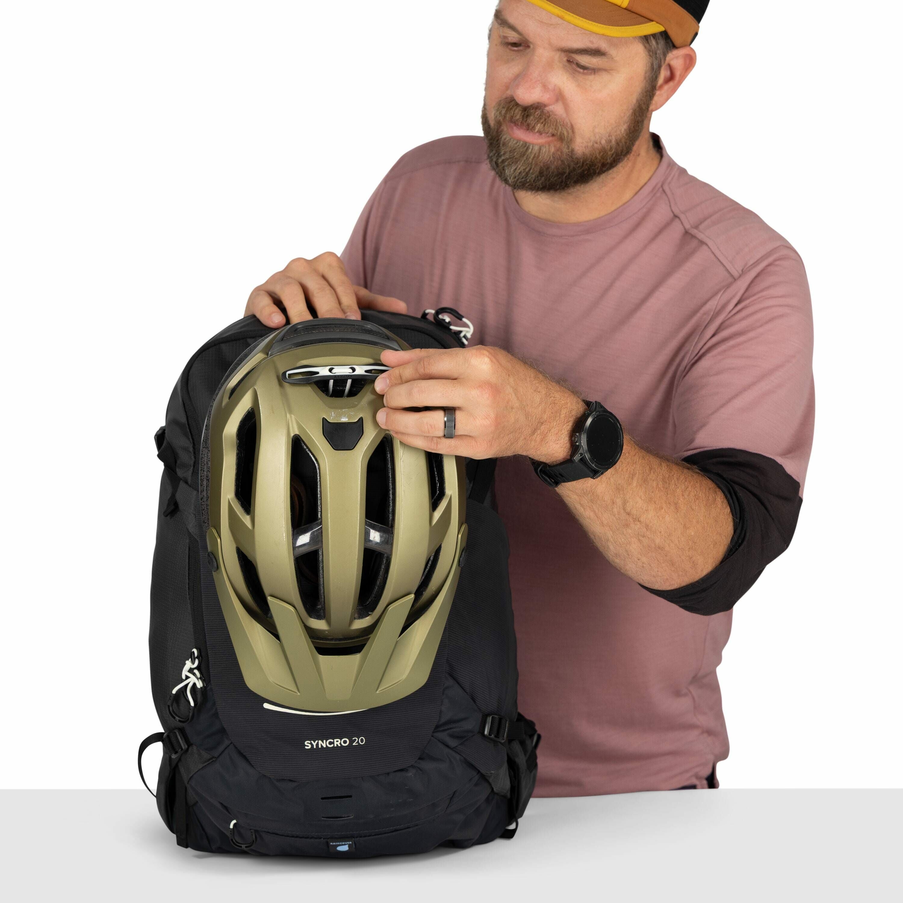 The image depicts a person wearing a pink long-sleeve shirt over a black undershirt, who is adjusting a helmet on a black backpack labeled "SYNCRO 20." The helmet is a light olive color with a logo "POC" visible on the front. The person is wearing a black