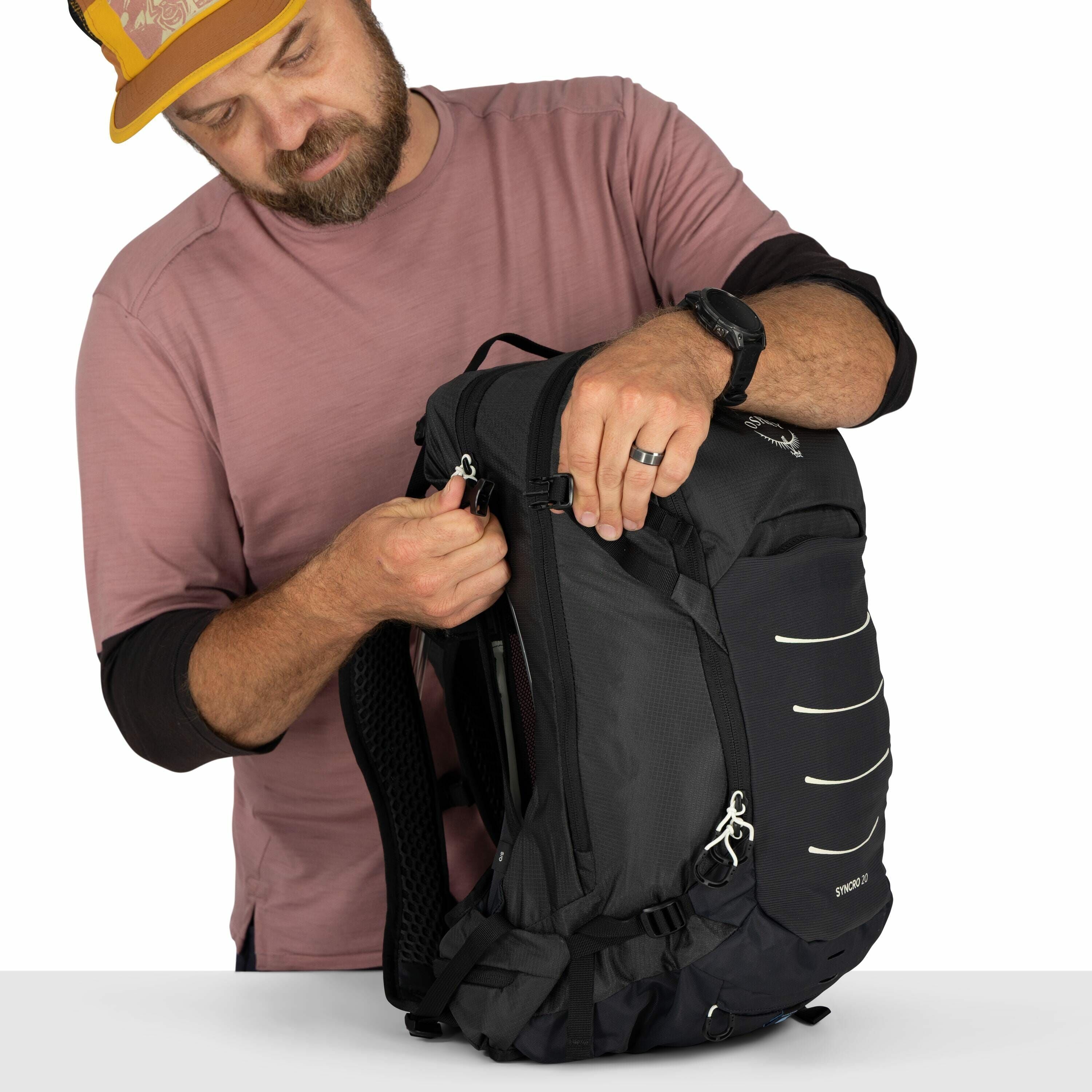 The image shows a person demonstrating the use of an Osprey Syncro 10 backpack. The individual is wearing a mauve long-sleeve shirt and a watch on the left wrist. They are in the process of adjusting or fastening a buckle on the side of the black backpack