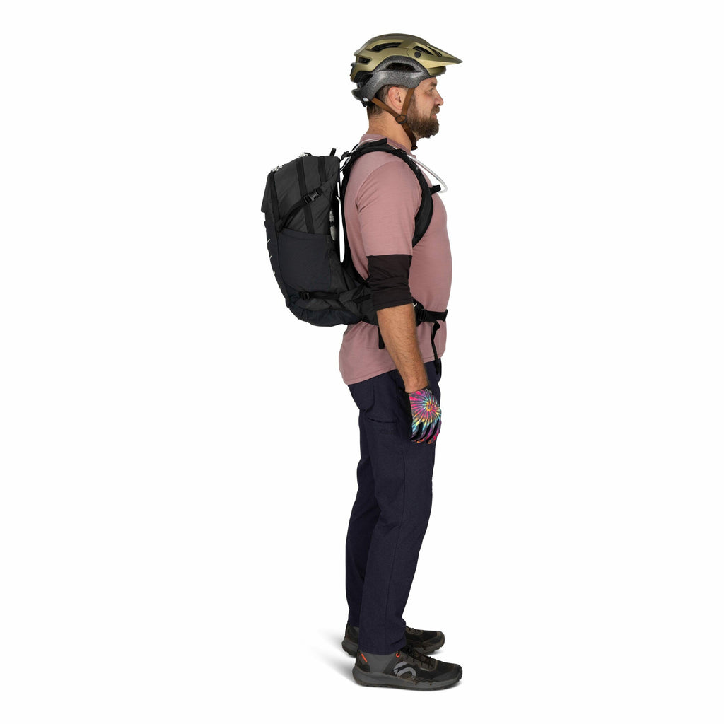 The image shows a man standing in profile wearing outdoor gear. He is dressed in a pink T-shirt layered over a long-sleeved black shirt, dark blue pants, and multicolored gloves. On his head, he wears a protective bicycle helmet, and he has a black backpa