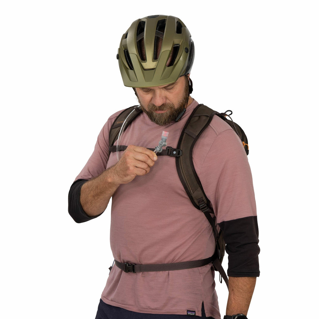 The image shows a person wearing a helmet, seemingly prepared for outdoor activities like cycling or hiking. They are dressed in a long-sleeve, layered top in a mauve color, with black sleeves visible underneath. The person is wearing a backpack with shou