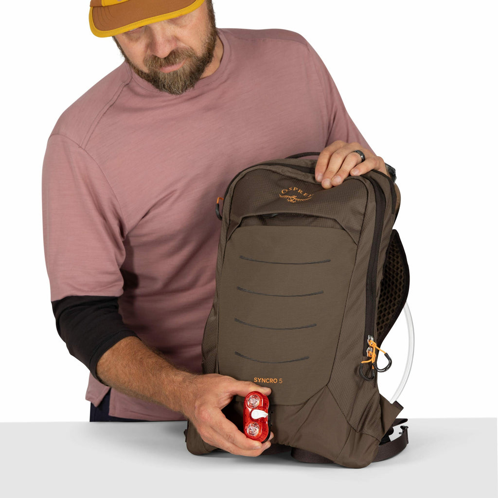 The image shows a person holding an Osprey Syncro 5 backpack, which is prominently displayed. The backpack is in a dark brown color and features several visible pockets and a streamlined design. The person is also holding a small red object, possibly a bi