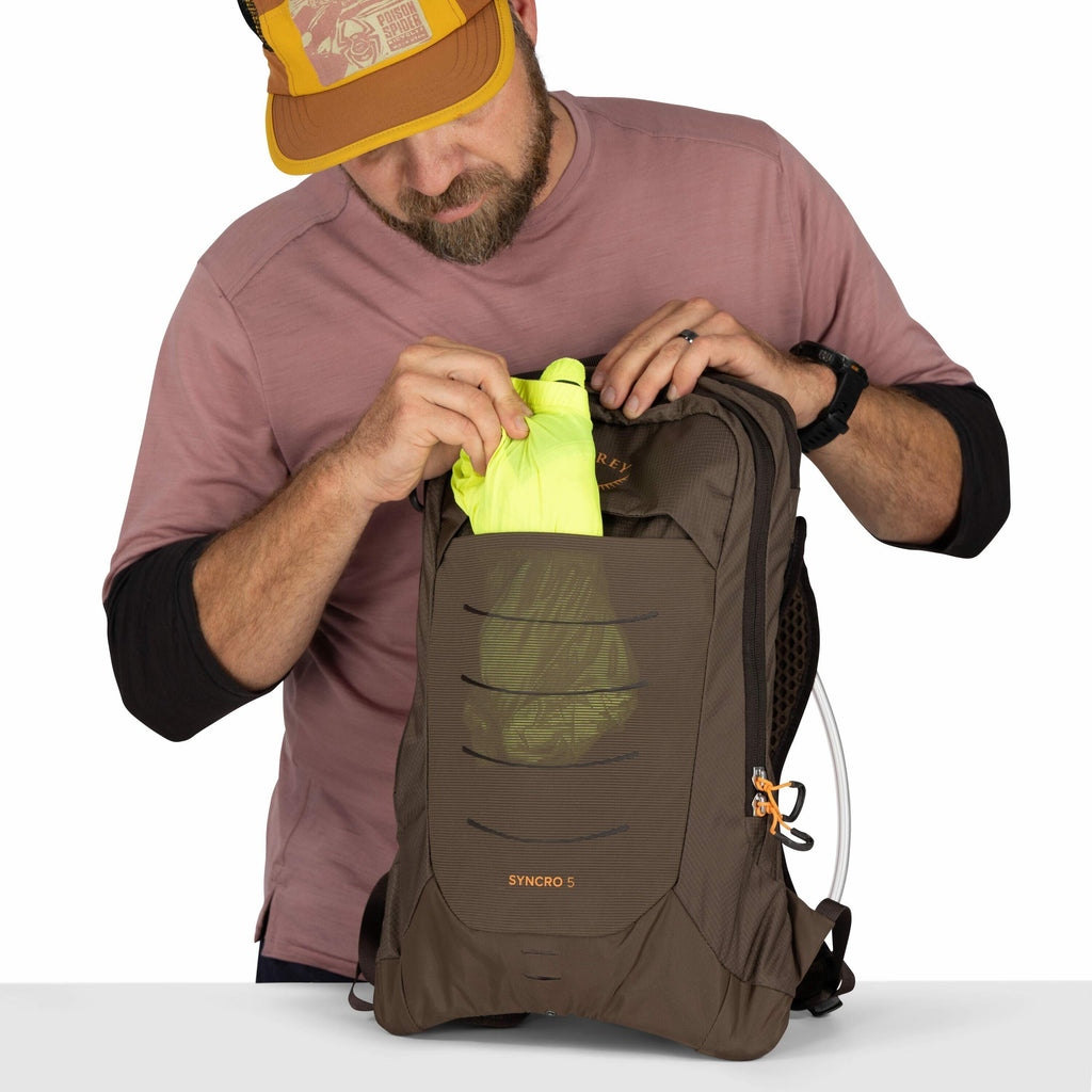 The image shows a person wearing a pink long-sleeve shirt and a yellow cap as they place a bright yellow garment into a brown backpack. The backpack appears to be an Osprey brand, as indicated by the visible logo and the model name "SYNCRO 5" printed in o