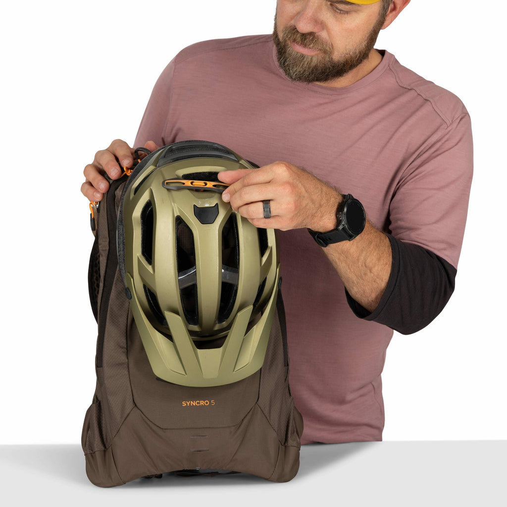 The image shows a man wearing a pinkish-brown long-sleeve shirt and a black smartwatch. He is holding a brown Osprey backpack, model "SYNCRO 5," to which an olive-green helmet is attached. The helmet is secured with a strap that has a small orange buckle.