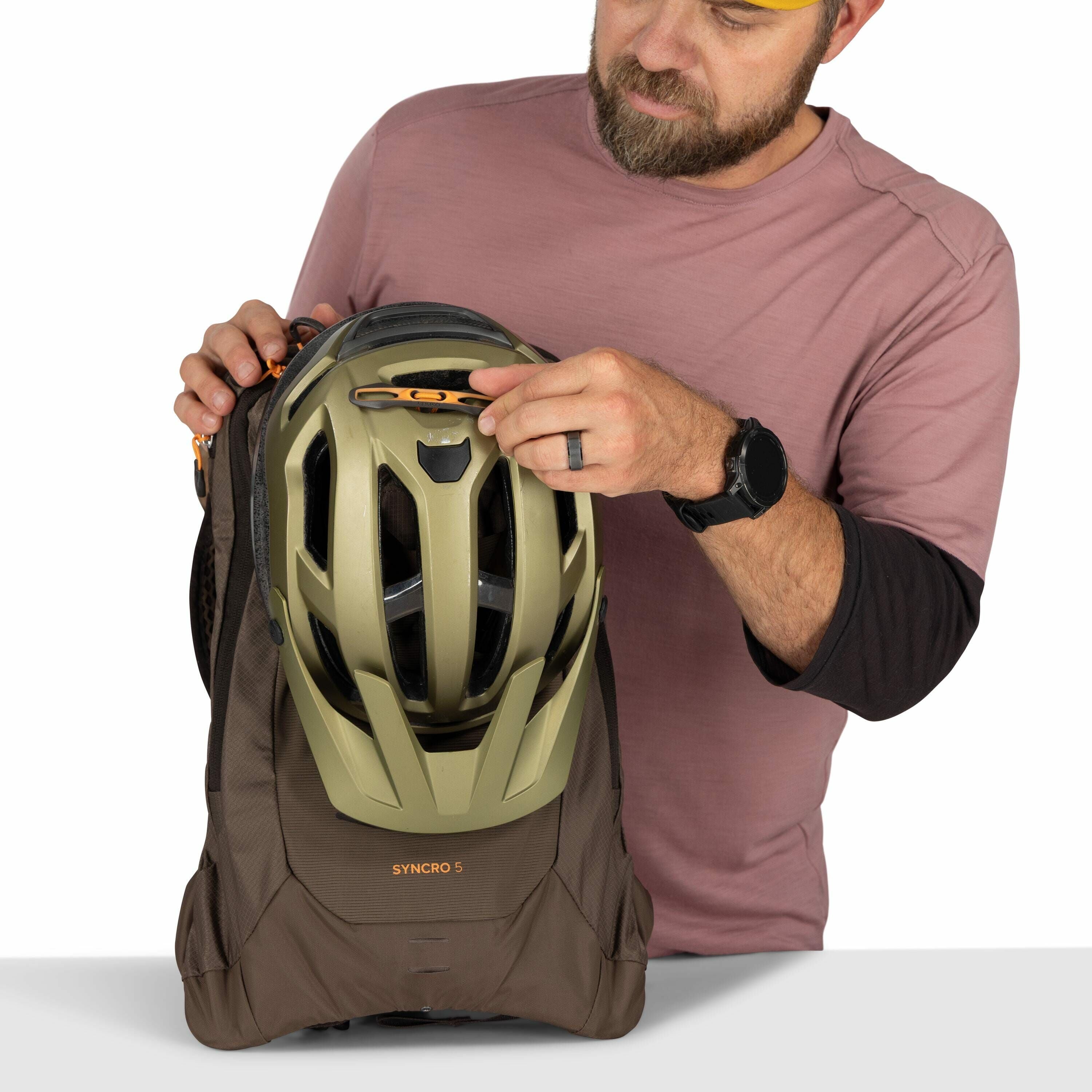 The image shows a man wearing a pinkish-brown long-sleeve shirt and a black smartwatch. He is holding a brown Osprey backpack, model "SYNCRO 5," to which an olive-green helmet is attached. The helmet is secured with a strap that has a small orange buckle.