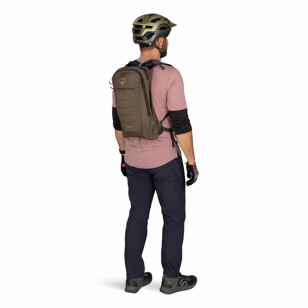 The image shows a man dressed in outdoor gear, likely prepared for cycling or hiking. He is wearing a helmet and a pair of gloves, with a long-sleeved pink shirt and dark pants. On his back, he carries a brown Osprey backpack, which seems to be designed f