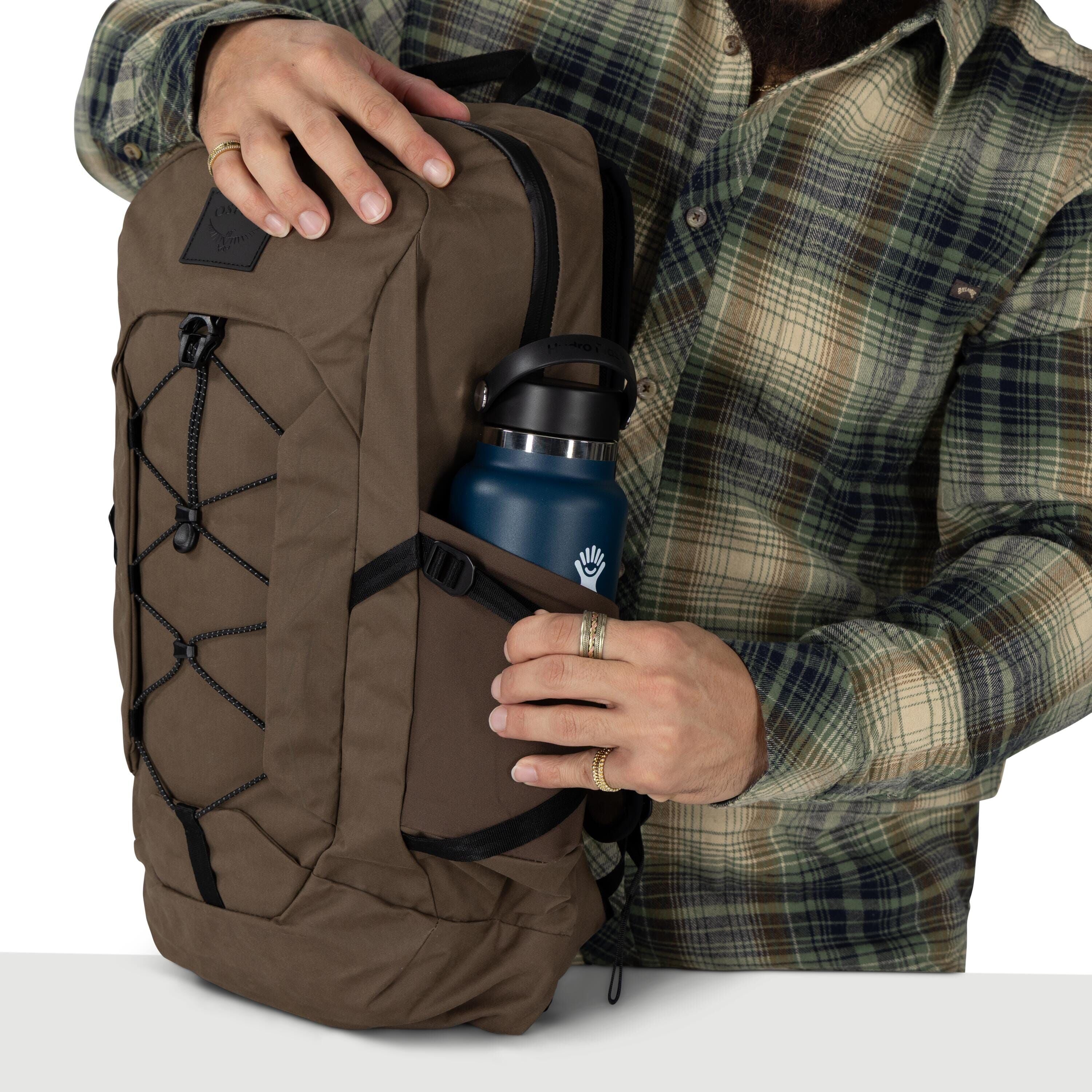 A person wearing a green plaid flannel shirt is holding a brown backpack. The pack has black bungee cords on the front and a side pocket that holds a blue Hydro Flask bottle with a black lid. The person?s hands, with a few rings, are adjusting the bottle