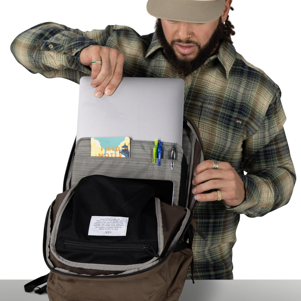 A person wearing a plaid shirt opens a backpack and slides a laptop into a padded compartment. The bag?s organizer panel shows a small ?Field Notes? notebook and several pens clipped in place. Another tablet or slim device is visible in a lower sleeve, an