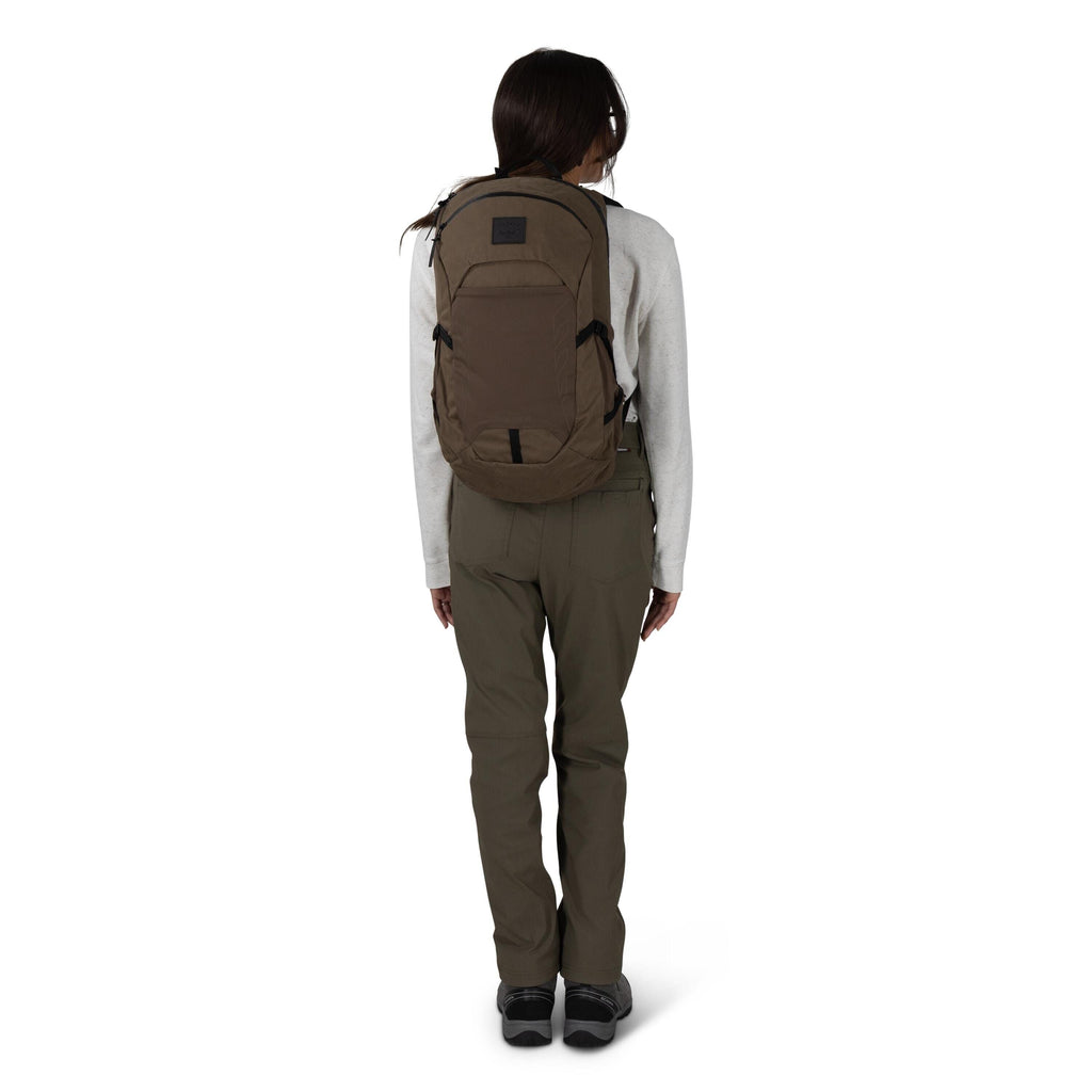 A person is standing with their back to the camera, wearing a khaki-brown backpack. They have on an off-white long-sleeve top, olive-green pants, and black shoes. The backpack has a streamlined shape with compression straps and a small patch logo near the