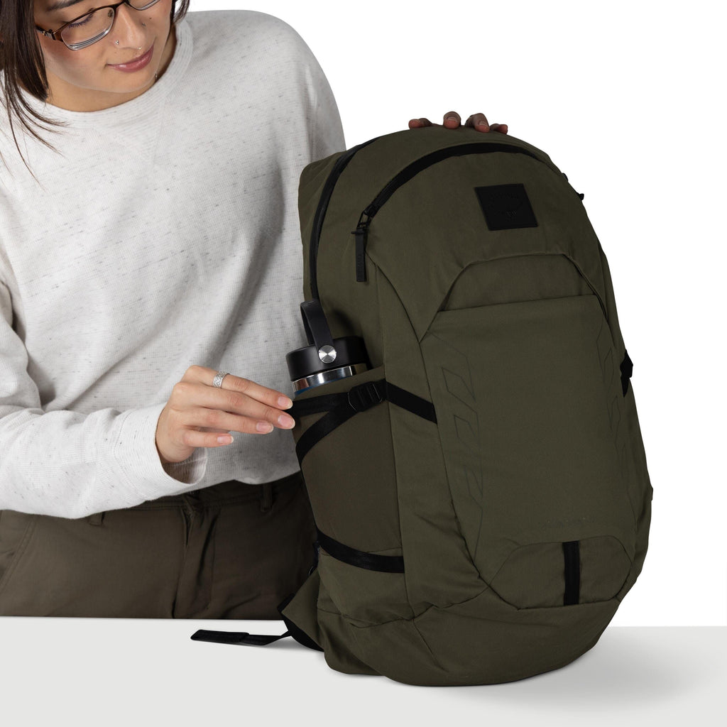 A person wearing glasses and a light gray sweatshirt is handling an olive green backpack. They are sliding a stainless steel water bottle into the side stretch pocket, which is secured by a black strap. The backpack has multiple zippered compartments, bla