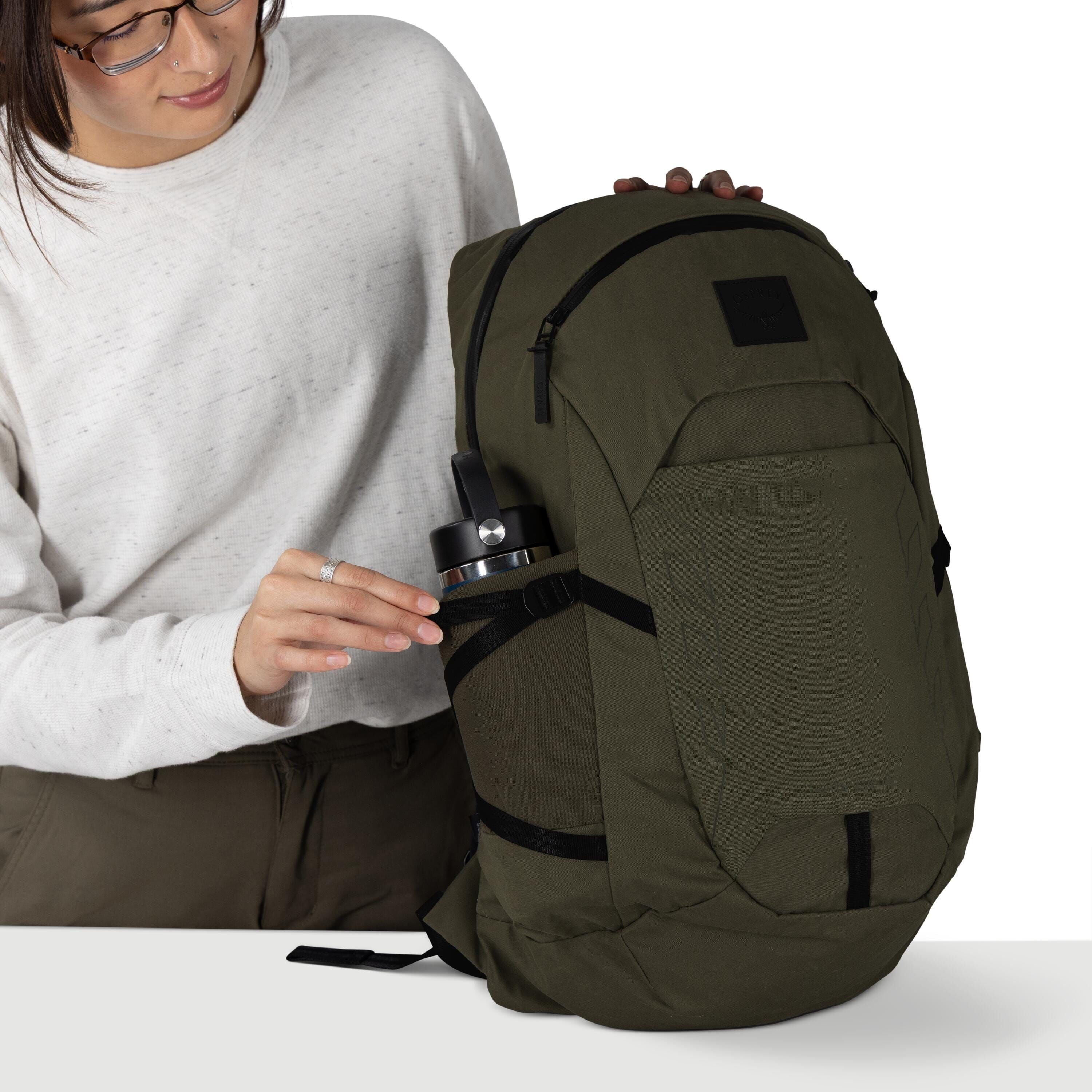 A person wearing glasses and a light gray sweatshirt is handling an olive green backpack. They are sliding a stainless steel water bottle into the side stretch pocket, which is secured by a black strap. The backpack has multiple zippered compartments, bla