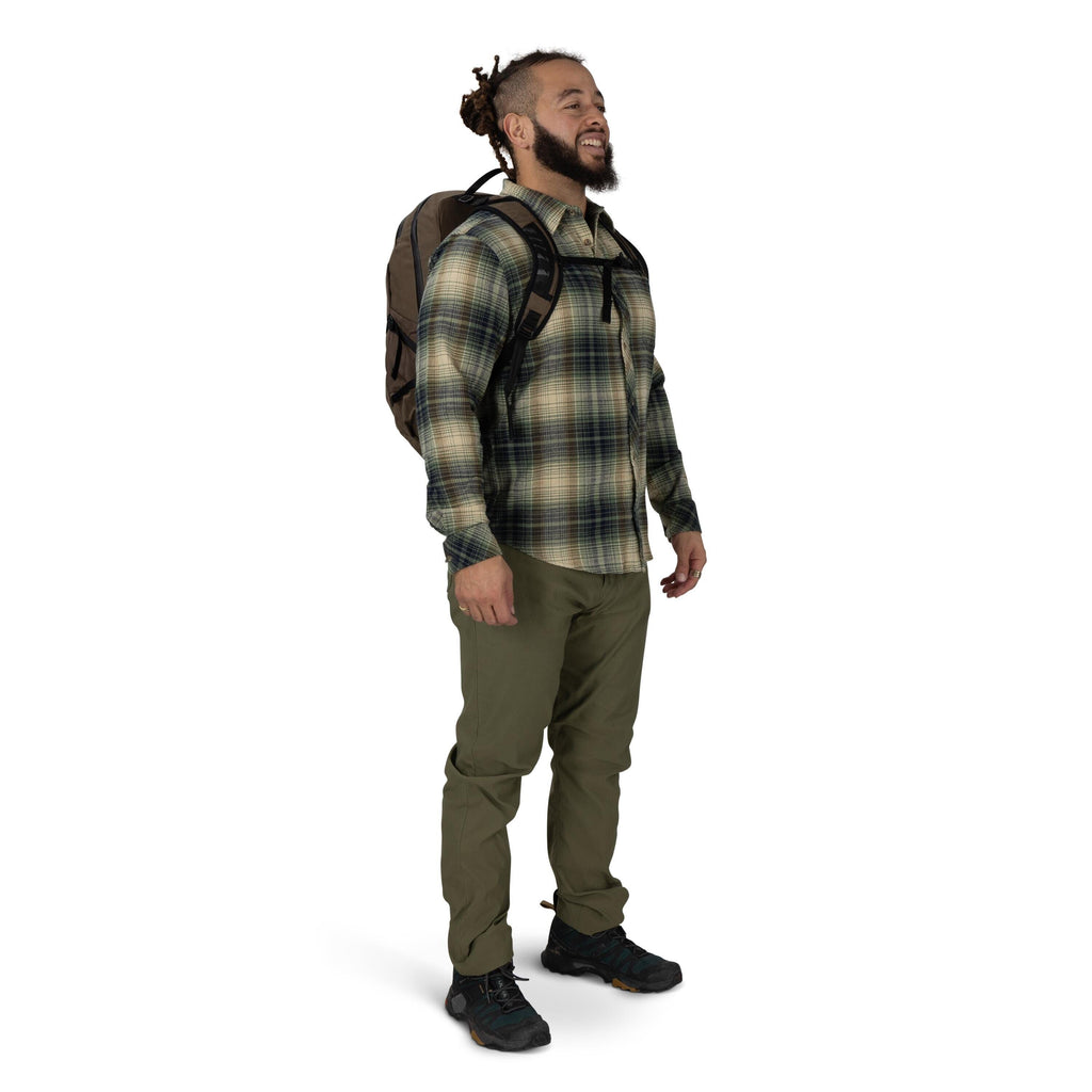 A person stands against a plain white background wearing outdoor clothing and a backpack. They have on a green and beige plaid button-up shirt, olive hiking pants, and black trail shoes. The backpack is brown with black straps, and a chest strap is clippe